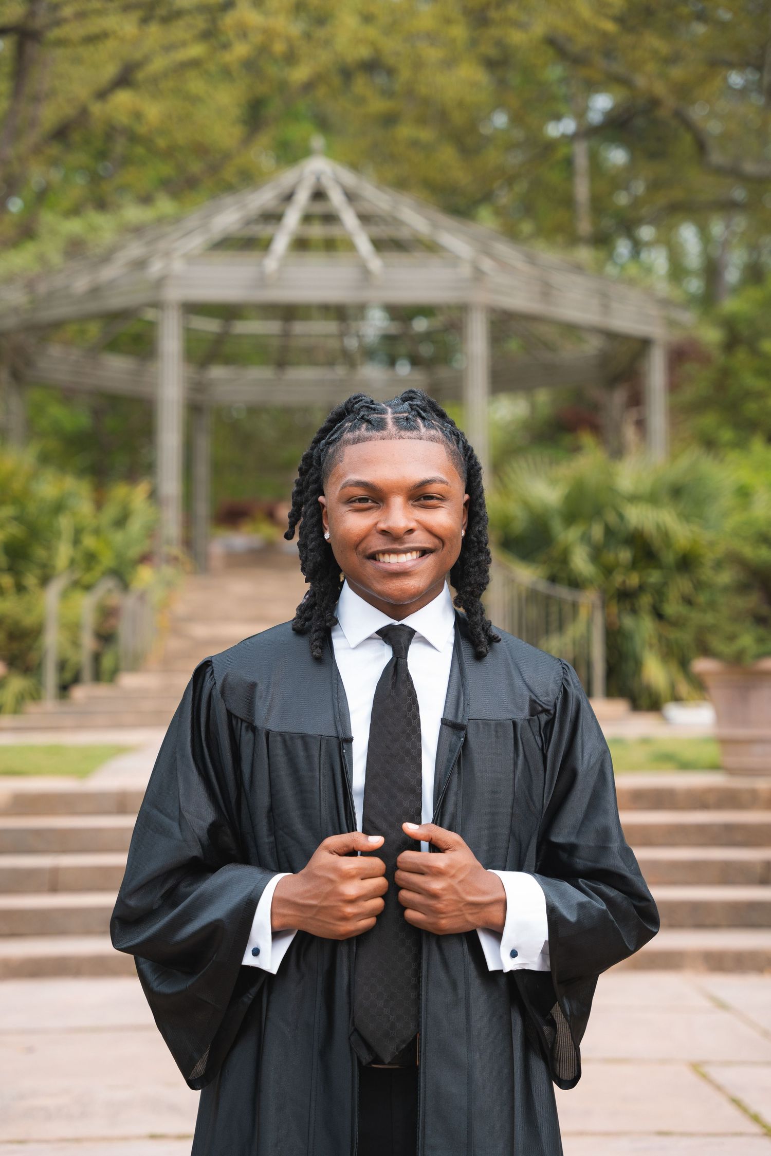 Graduate in a black gown standing on garden steps in front of a wooden pergola
