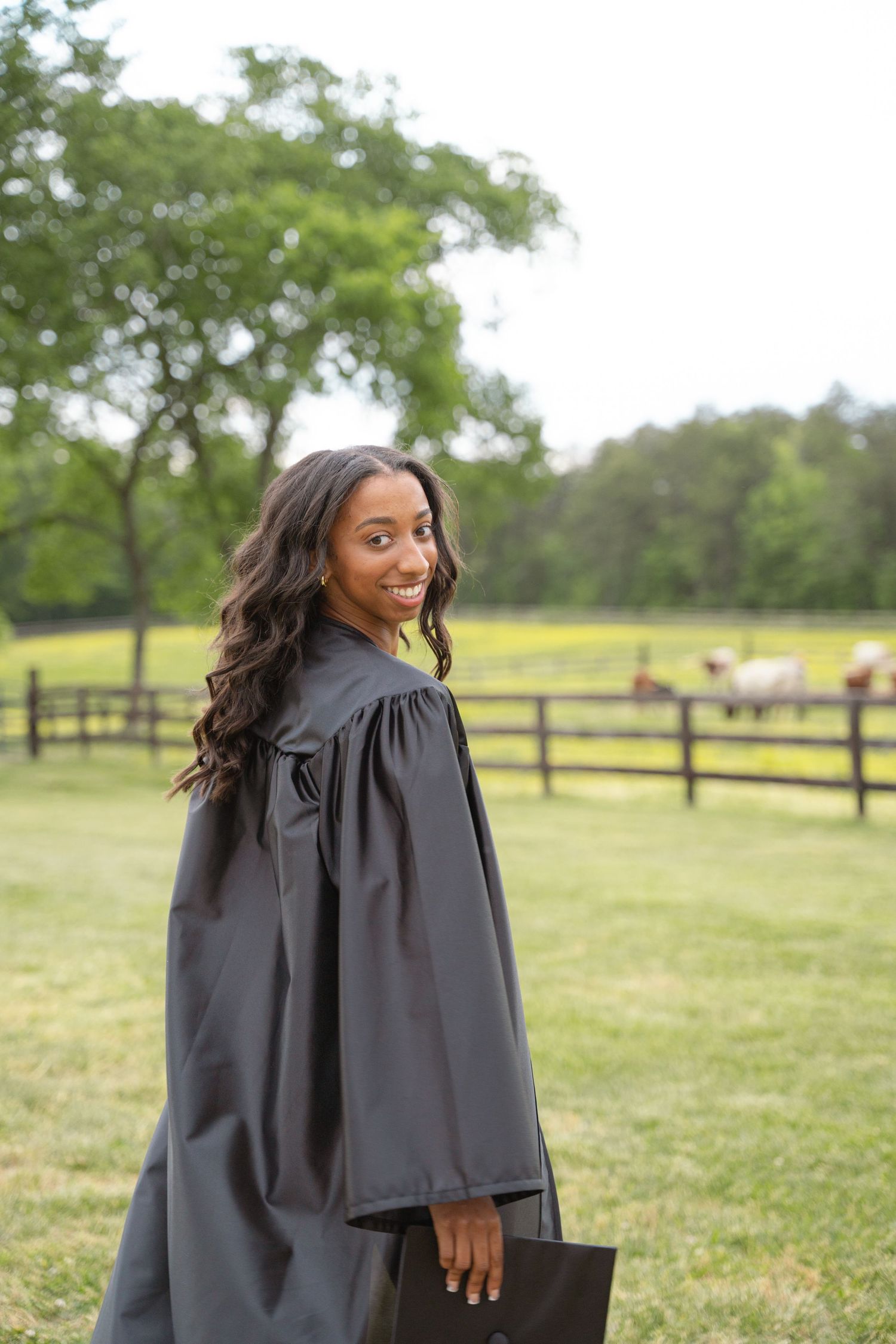 Graduate in cap and gown standing in a field with a split-rail fence behind her