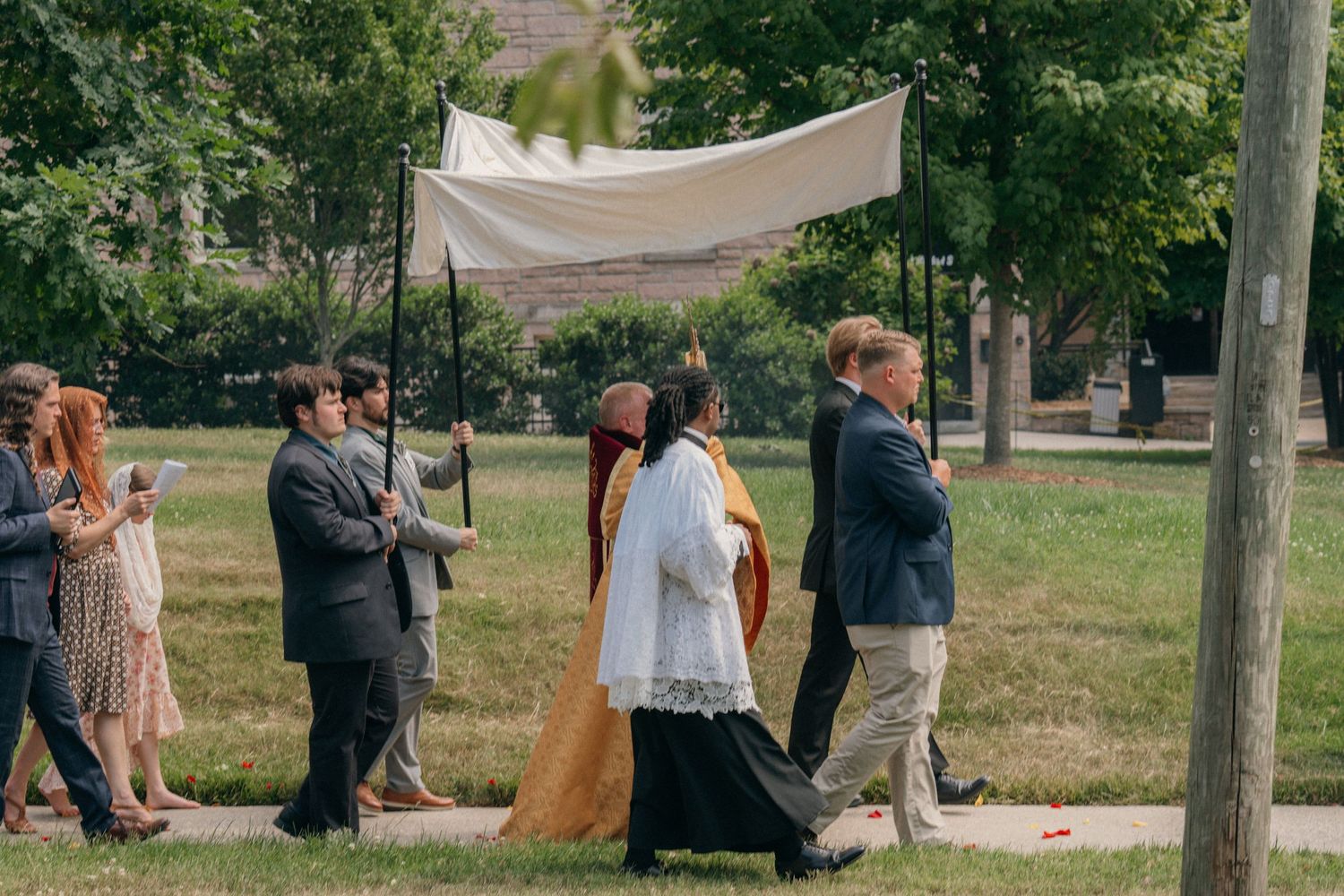 Priest carrying the Eucharist under a white canopy in a Corpus Christi procession on a church lawn