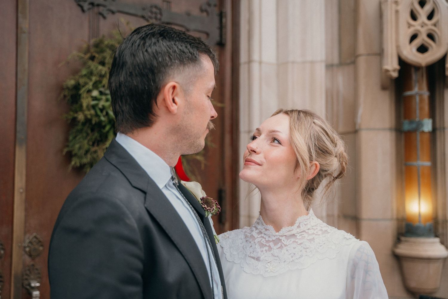 Groom and bride smiling at each other beside the carved stone church doorway