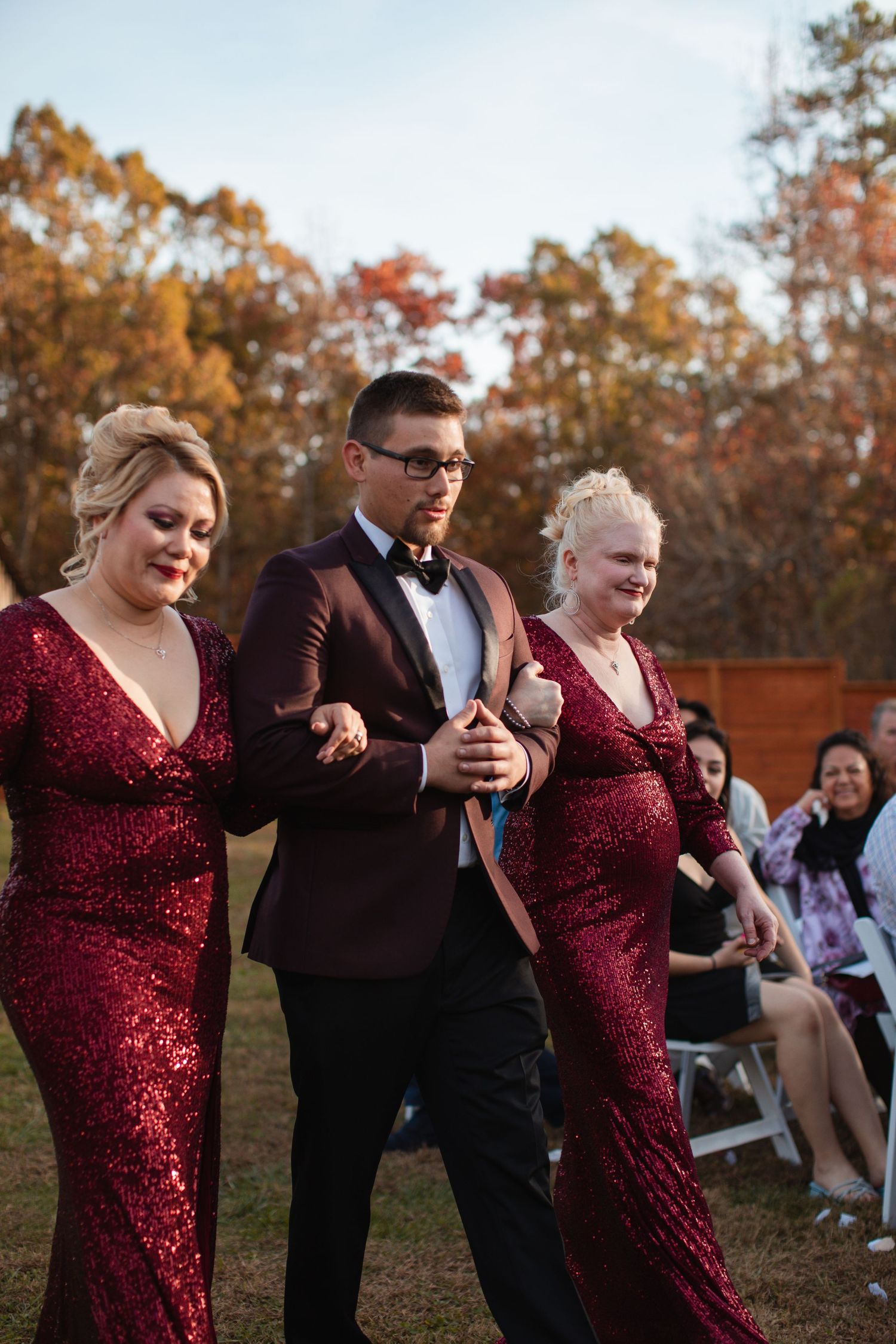 Groom escorting bridesmaids in burgundy sequin dresses down the aisle