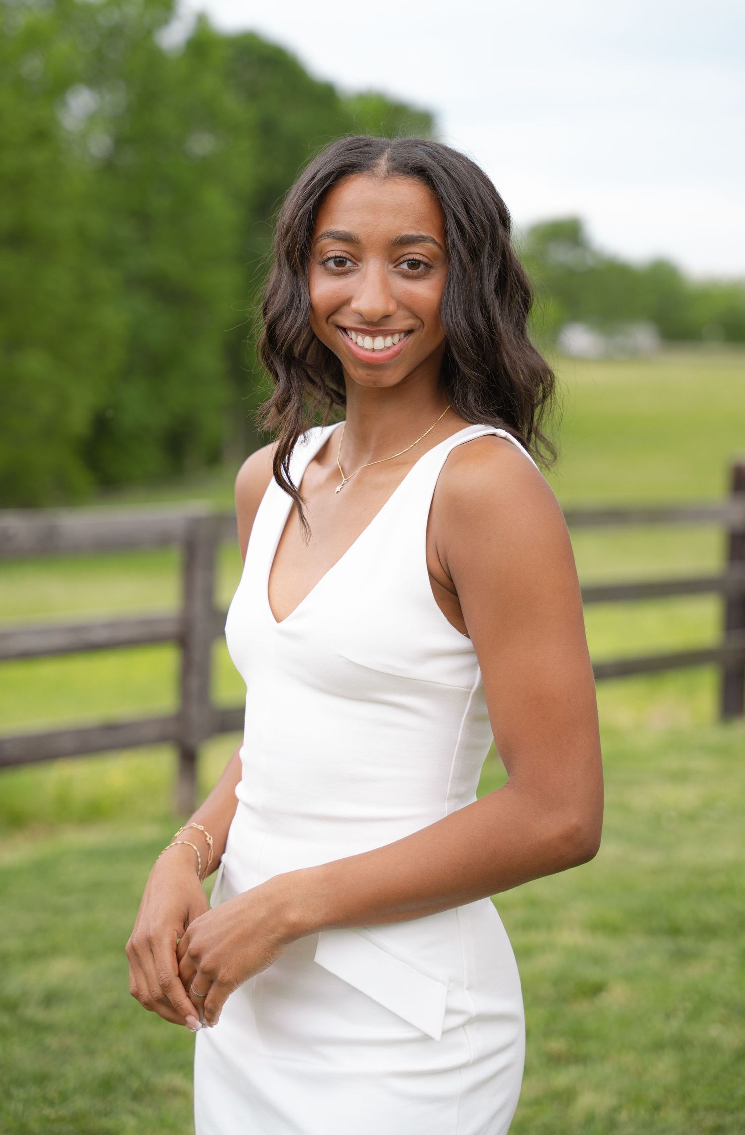 Smiling graduate in a white dress standing in a green pasture by a wooden fence