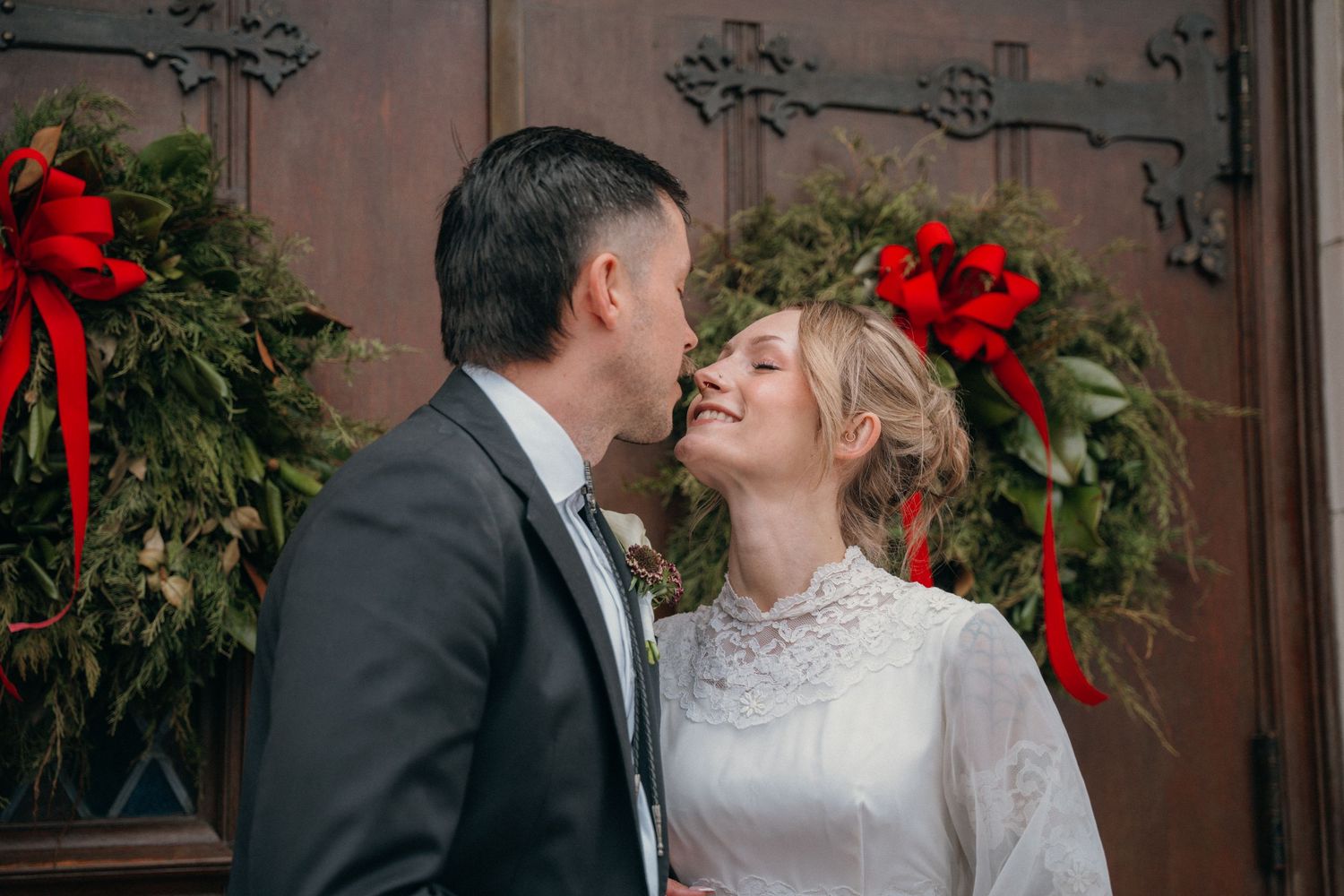 Groom kissing the bride's forehead in front of Christmas wreaths