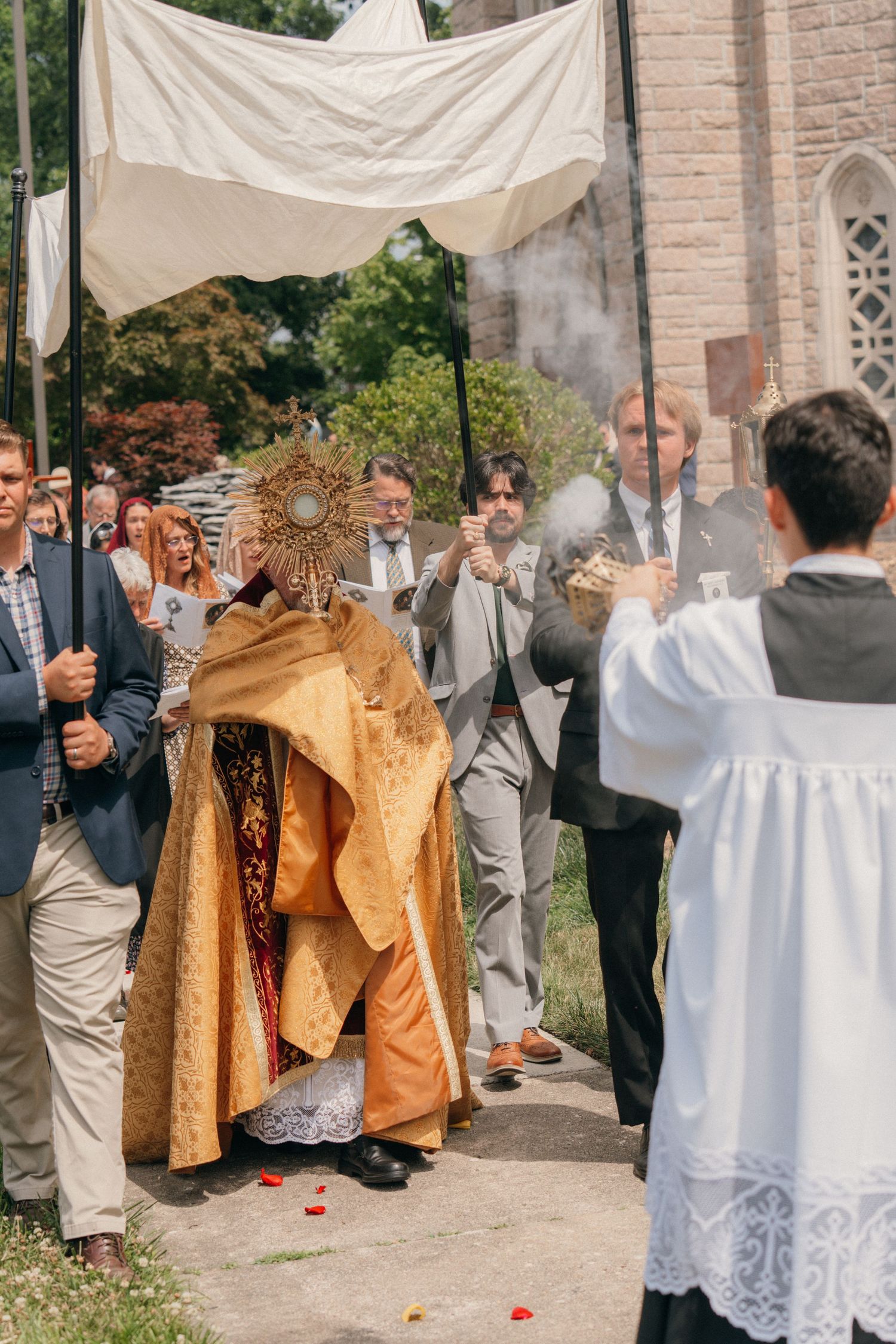 Priest pausing with the Blessed Sacrament under a canopy as incense rises during Corpus Christi