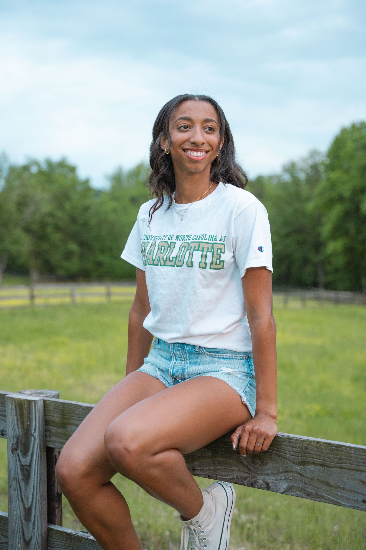 High school graduate in a college T-shirt and denim shorts sitting on a wooden farm fence