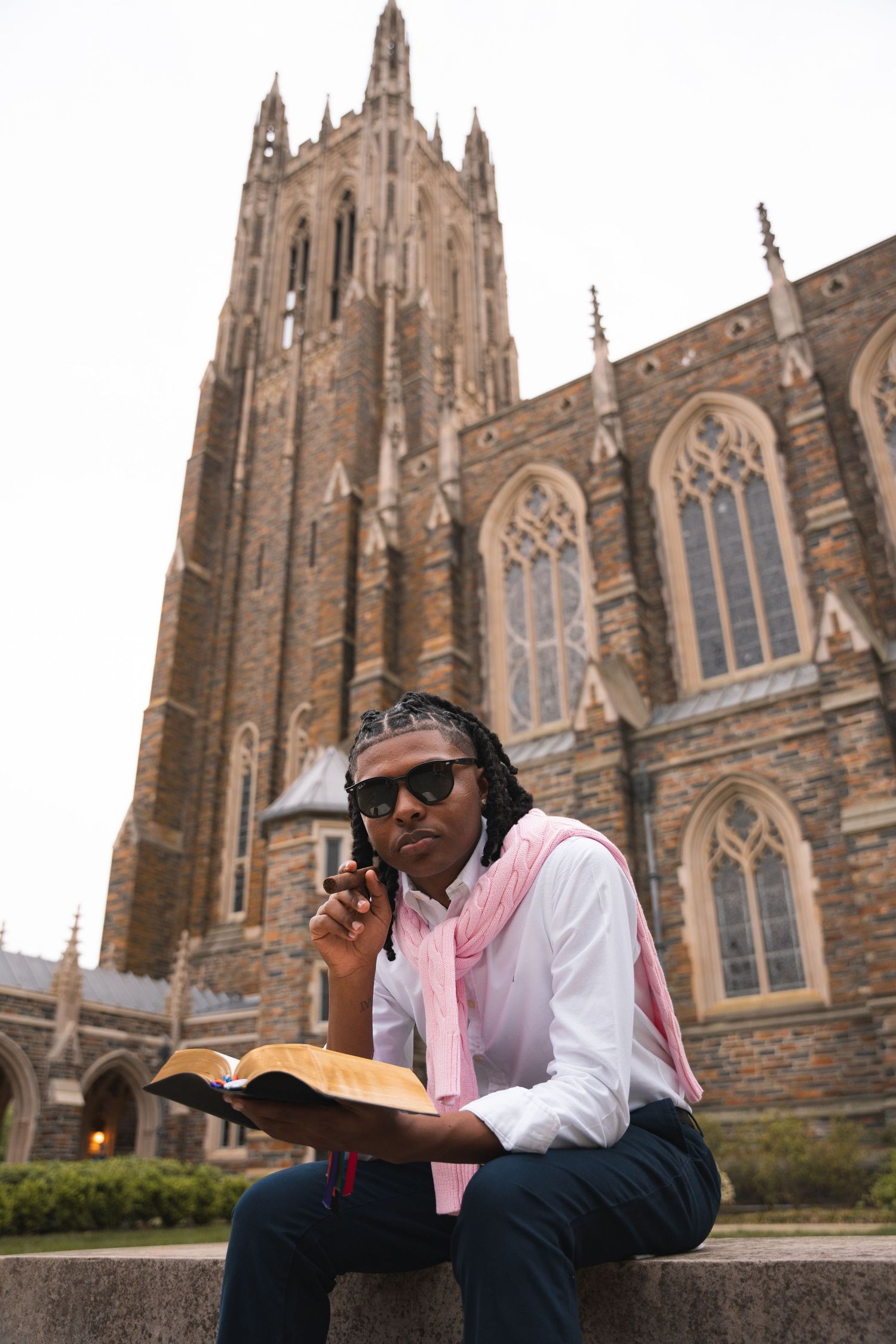 Graduate reading a Bible on a ledge beneath a tall Gothic chapel tower