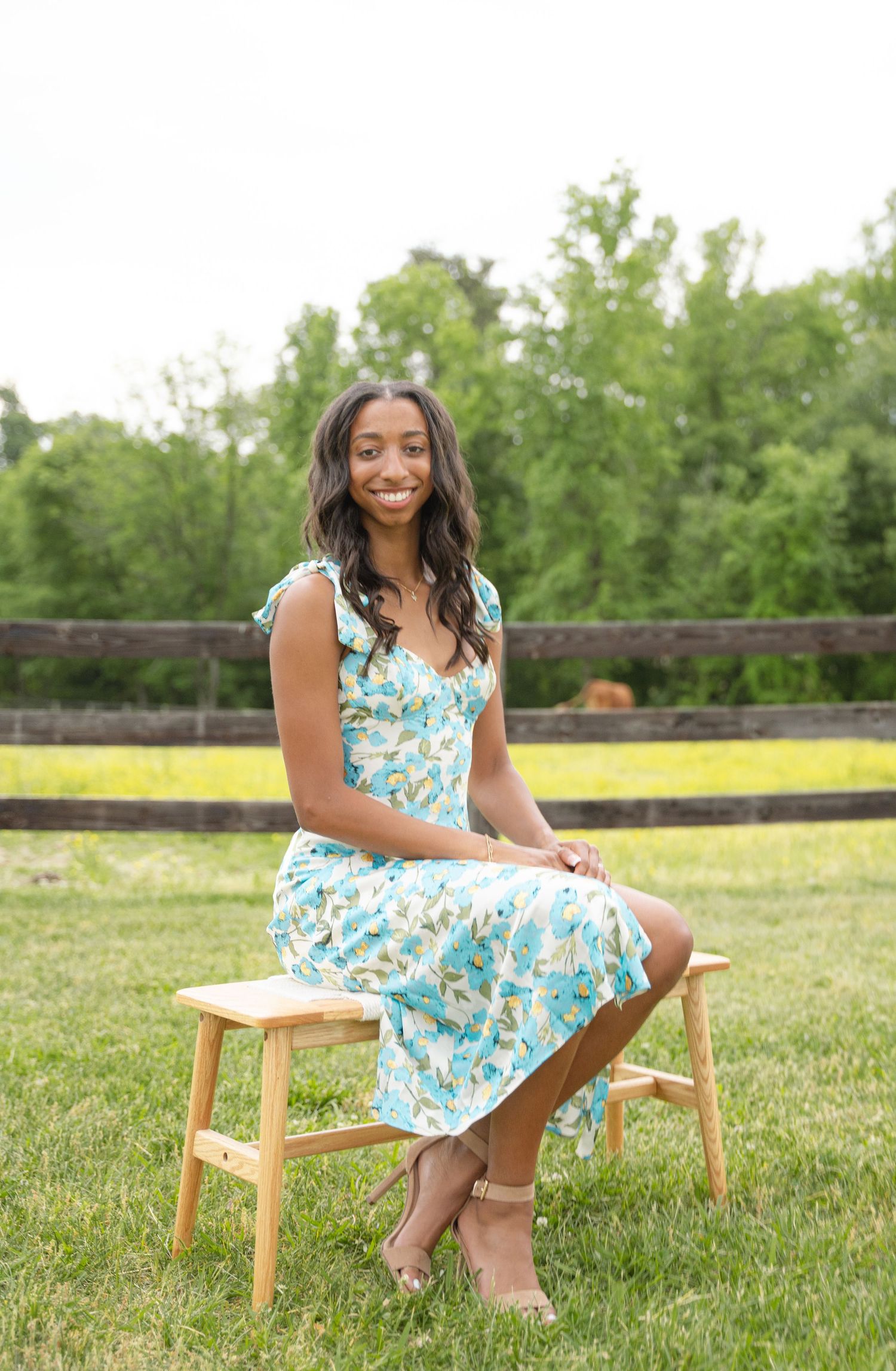 Graduate in a floral sundress posing on a wooden stool in a grassy meadow
