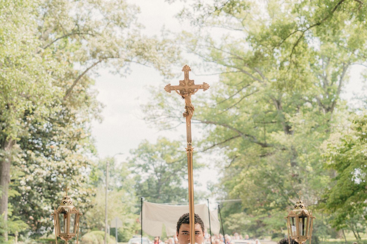 Processional cross leading a Corpus Christi procession beneath tall green trees