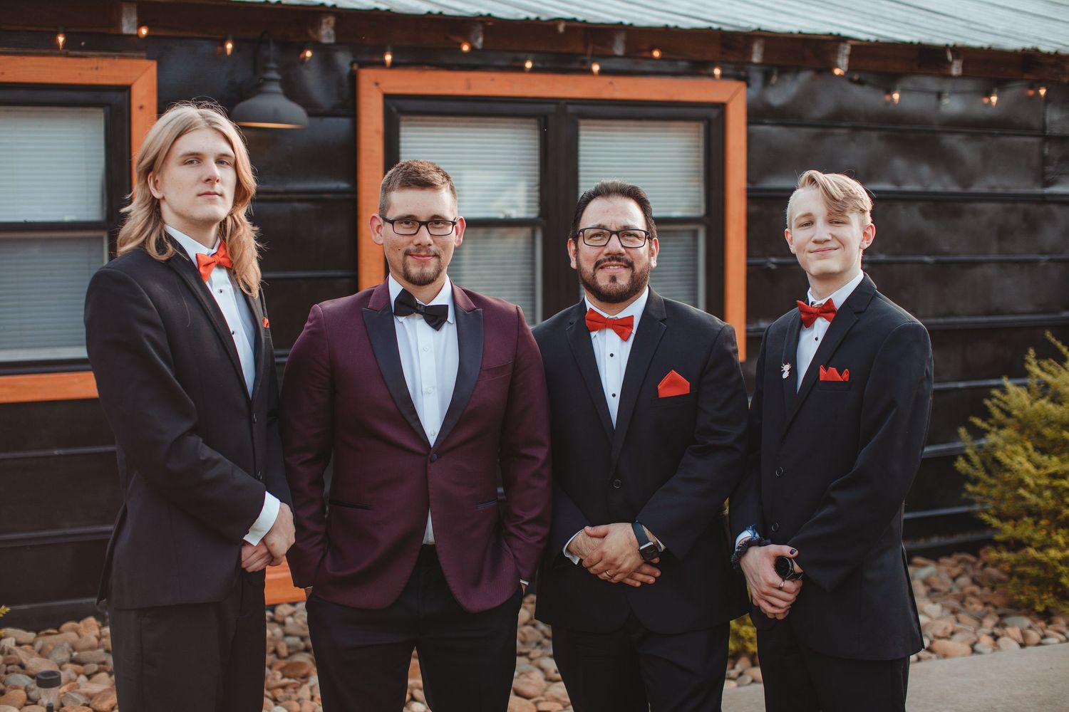 Groom and groomsmen in black suits and red bow ties at a rustic wedding venue