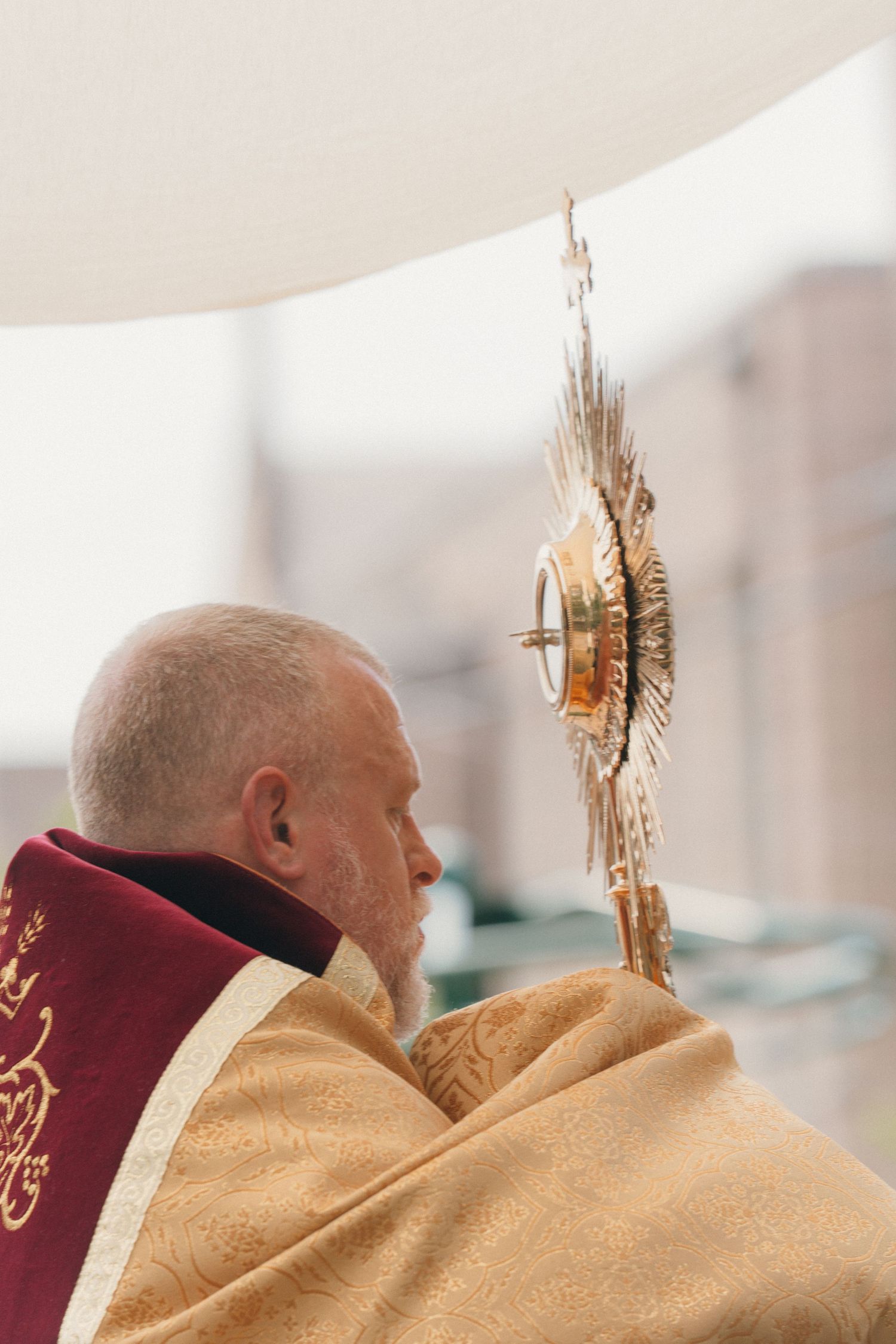 Priest in a gold humeral veil holding the monstrance under a canopy during a Corpus Christi procession