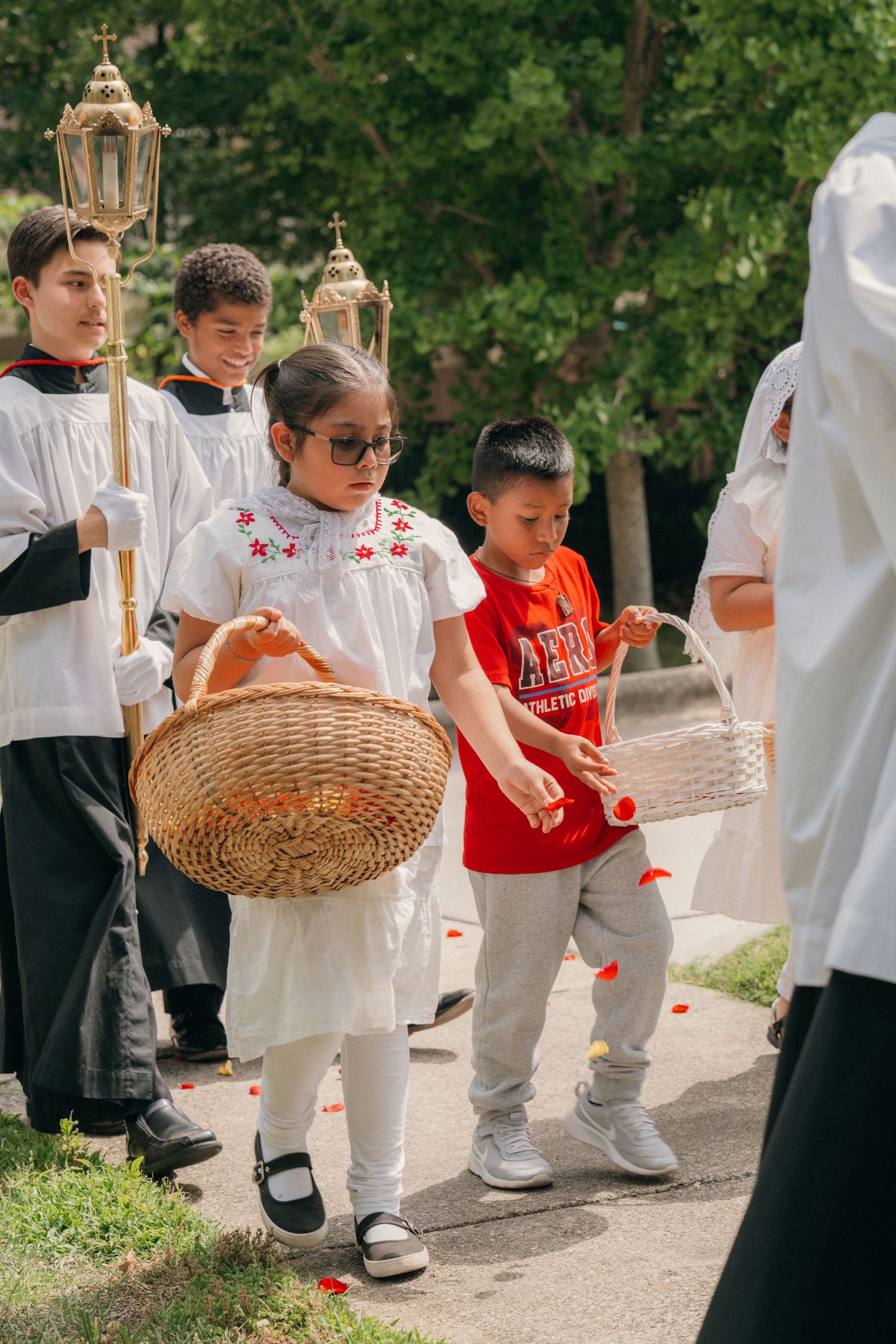 Children scattering flower petals from baskets ahead of a Corpus Christi procession with altar servers