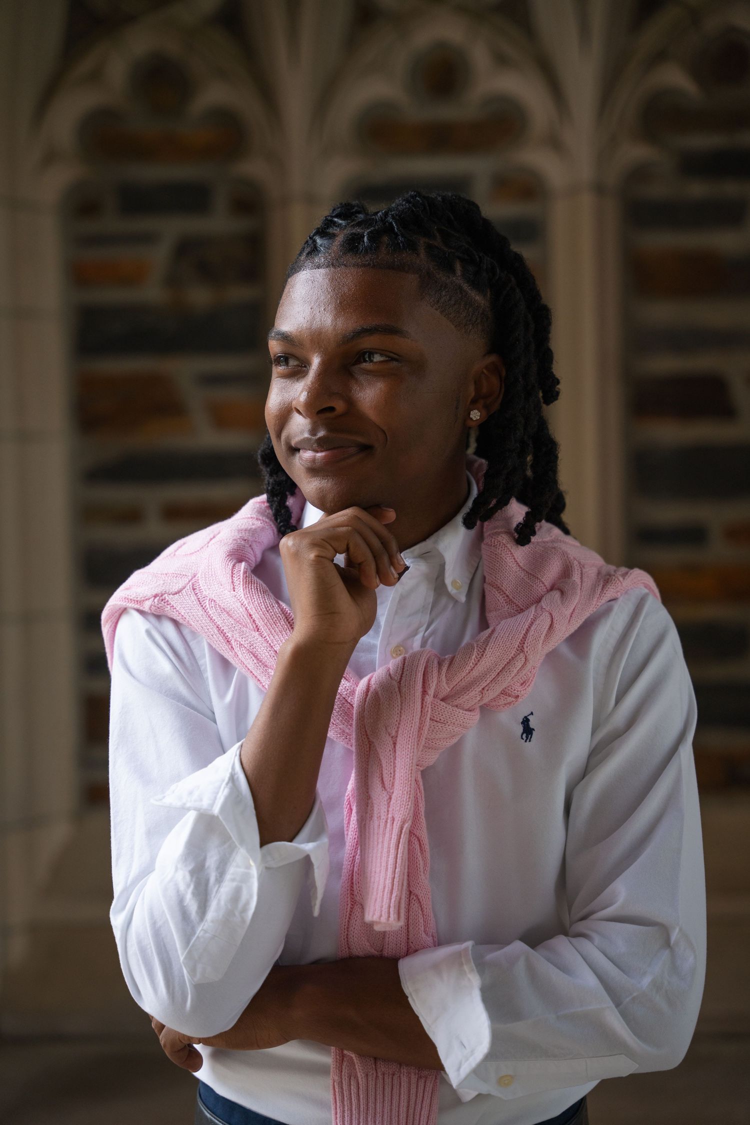 Graduate in a pink sweater with hand on chin under a stone archway