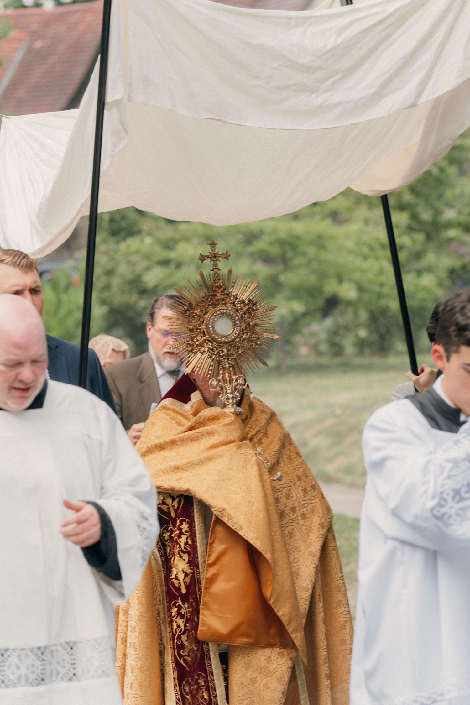 Priest carrying the Blessed Sacrament under a canopy flanked by altar servers in white surplices