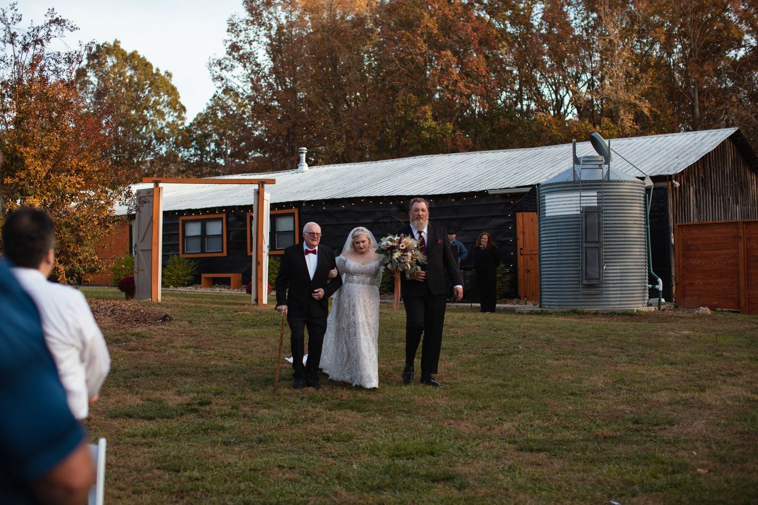 Bride escorted down a grassy aisle toward a rustic wedding venue