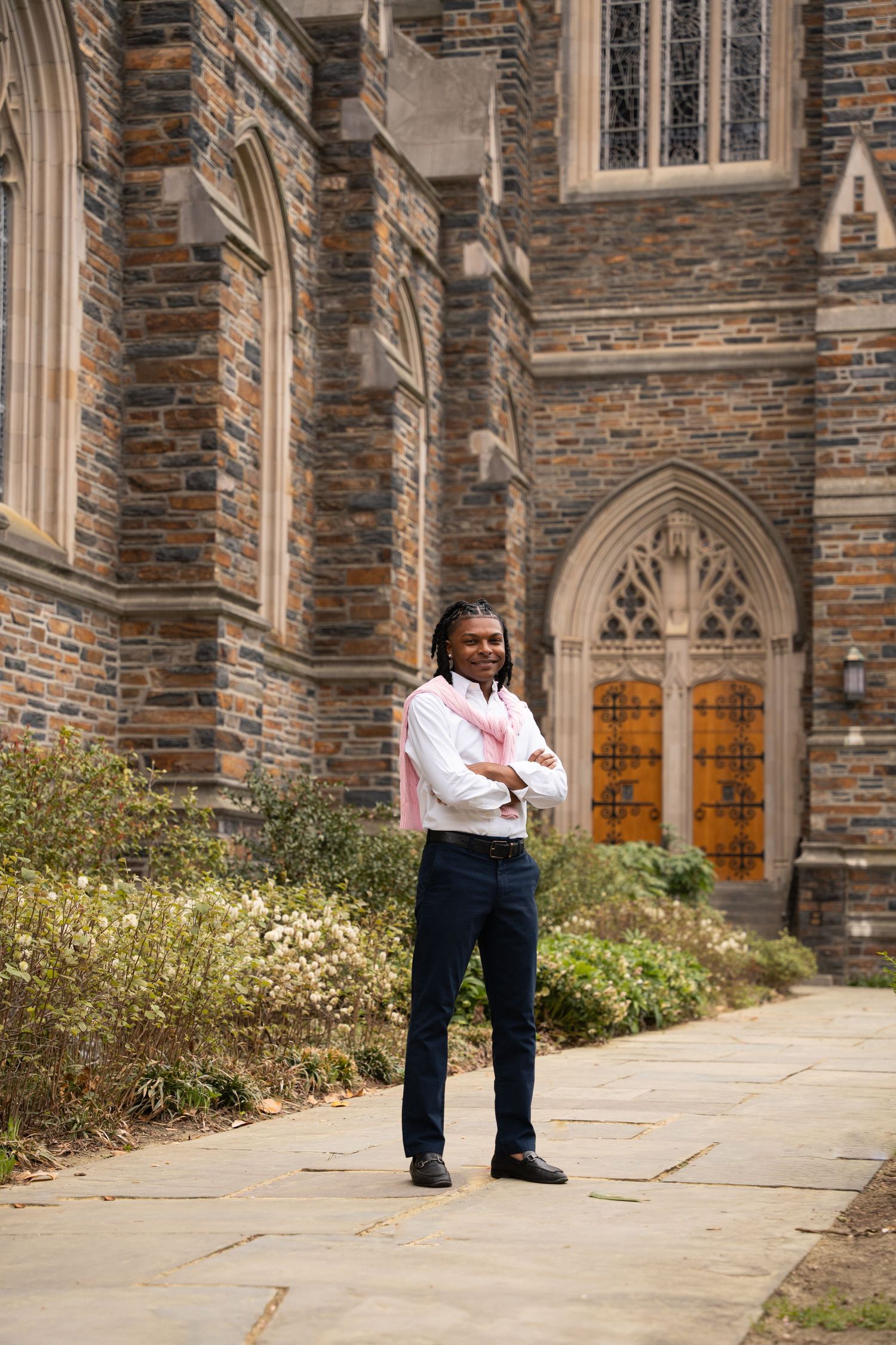 Graduate standing on a stone walkway outside a Gothic chapel framed by gardens