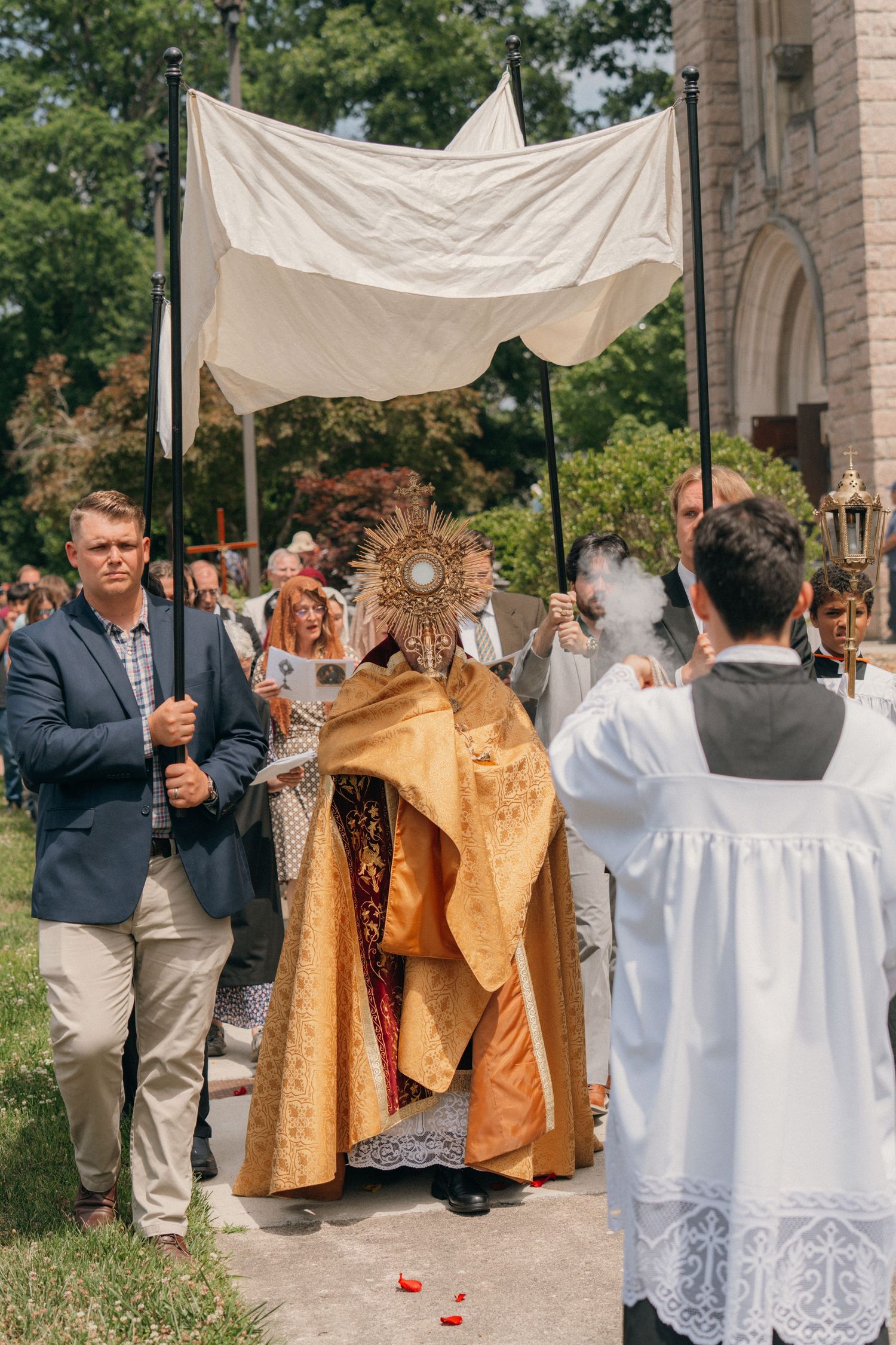 Priest holding the monstrance under a canopy as altar servers kneel nearby on Corpus Christi