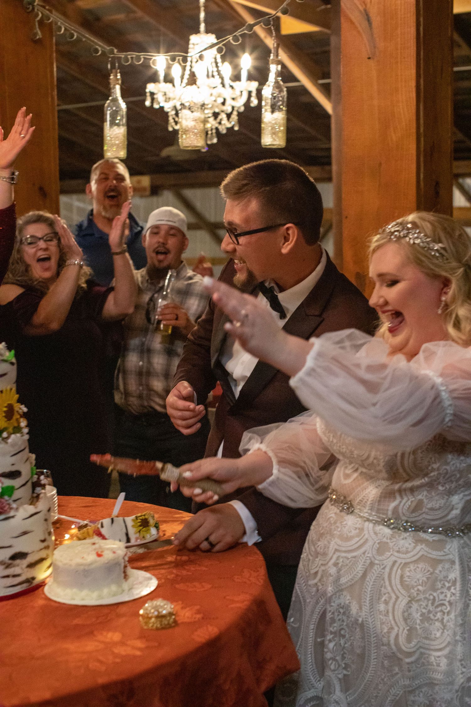 Bride and groom cutting their wedding cake as guests cheer beneath a chandelier