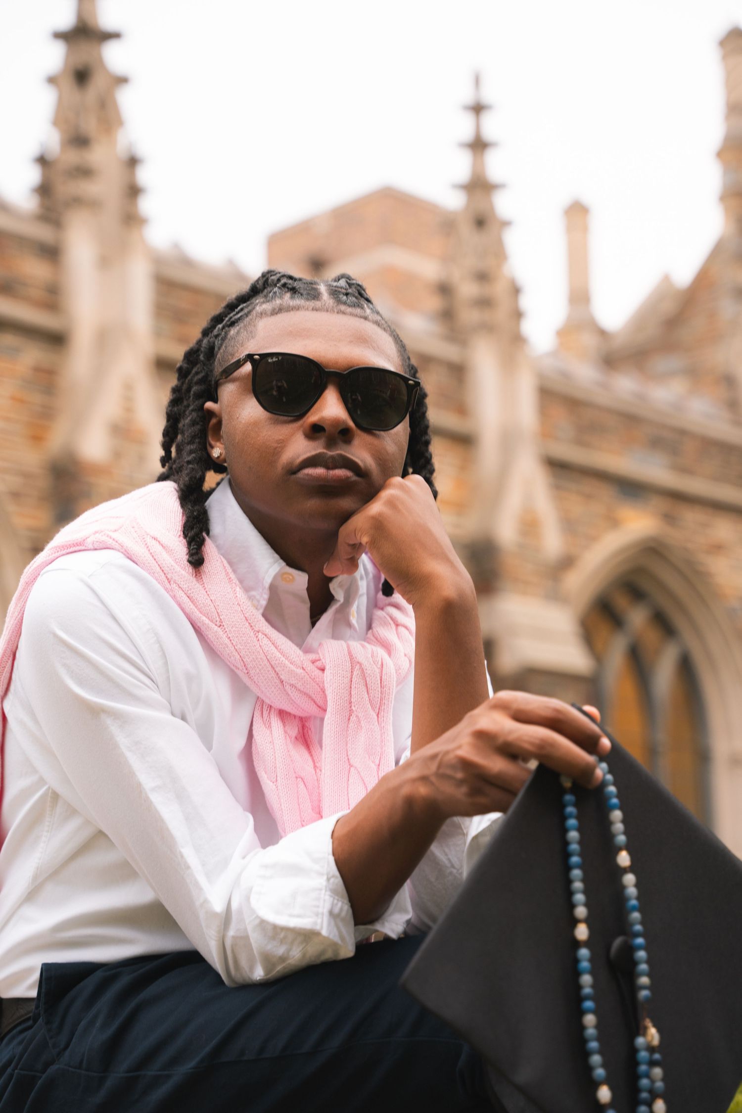 Graduate in sunglasses holding rosary beads and his cap outside a stone chapel