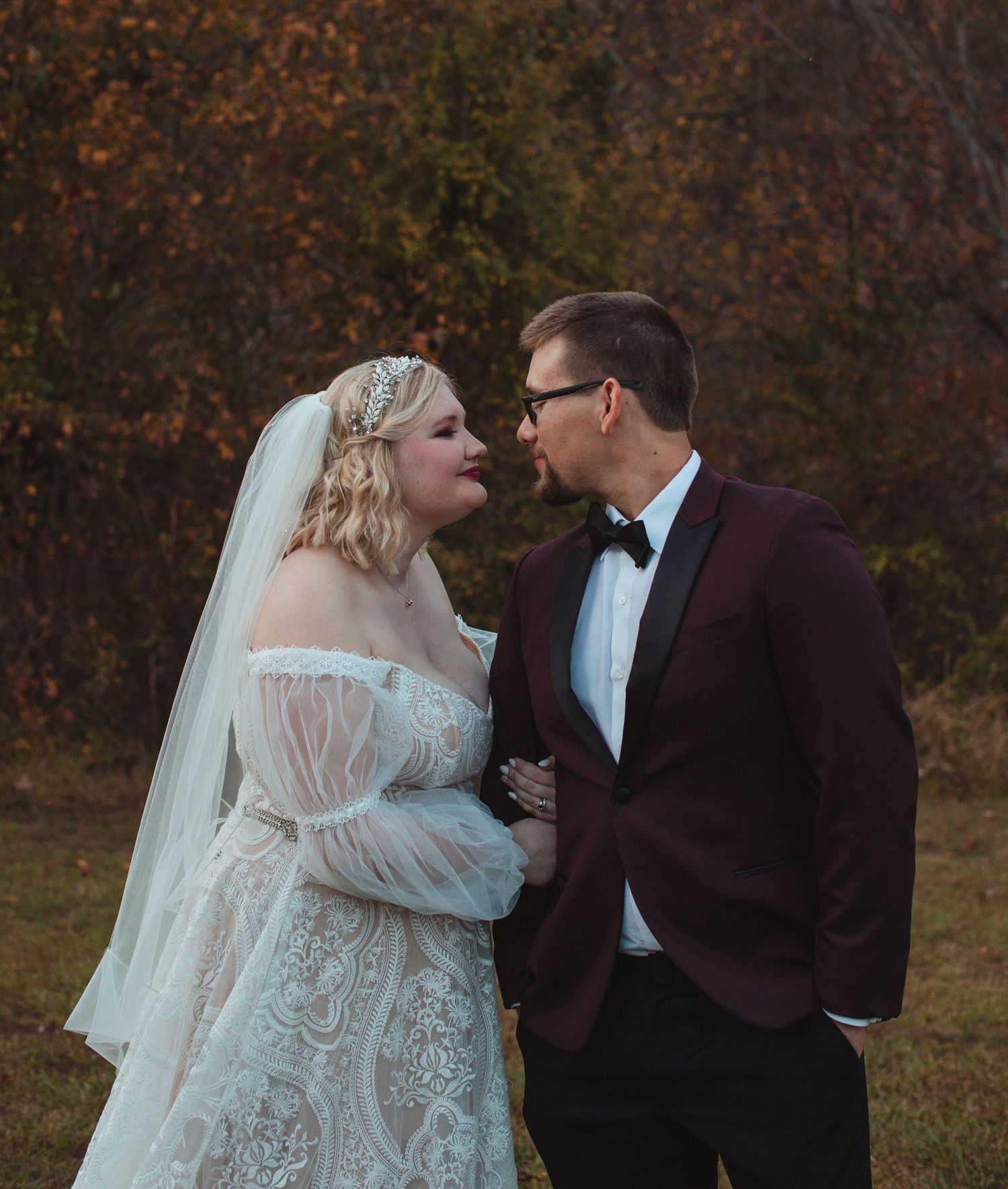 Bride in a lace gown and veil with the groom in a burgundy tuxedo jacket in autumn woods