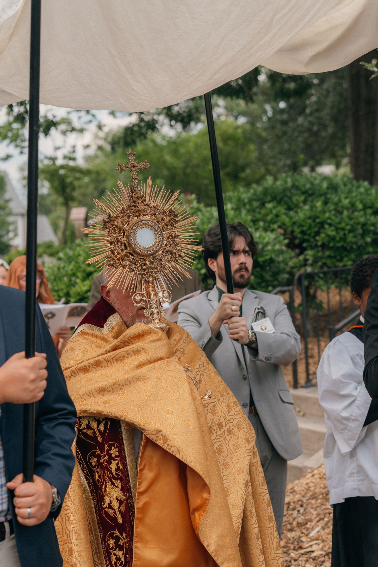 Priest in gold vestments carrying the monstrance, canopy bearers at his side, on Corpus Christi