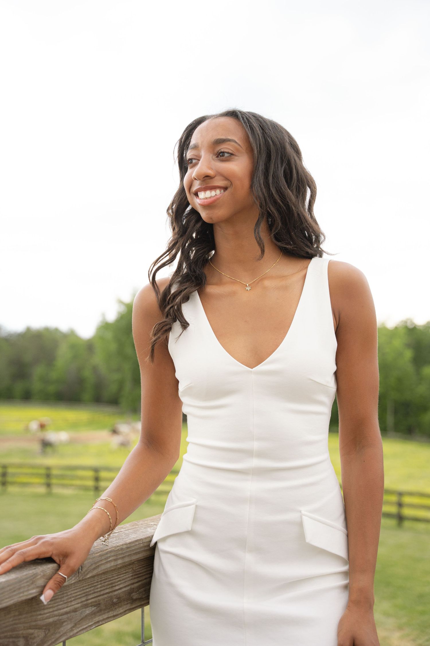 Graduate in a white dress smiling beside a wooden fence on a farm
