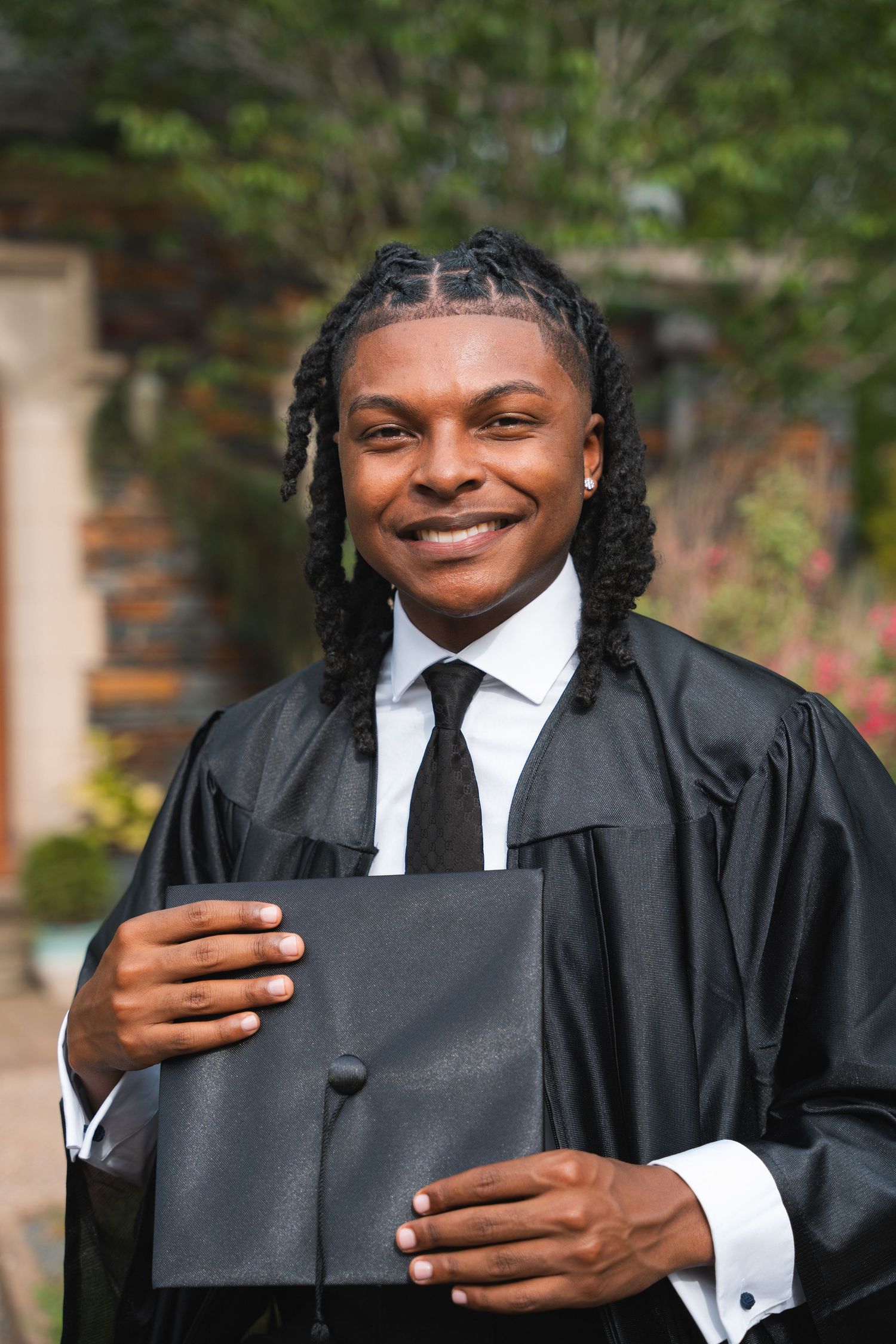 Smiling graduate in a black gown holding his cap in a garden