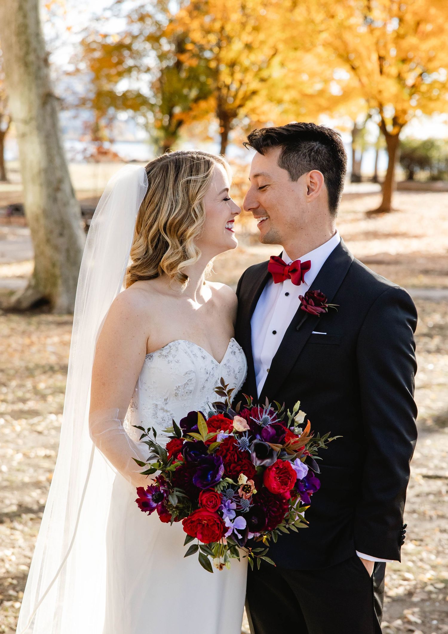 A wedding couple poses with a vibrant red and purple bouquet against an autumn backdrop. Philadelphia wedding. Switch House Cescaphe.