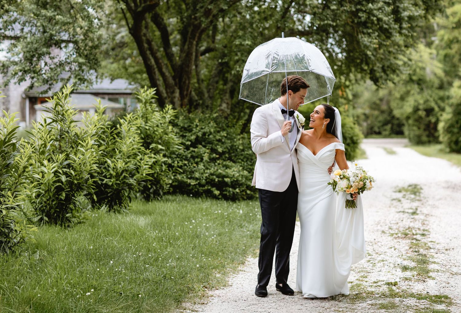 Wedding couple and photographer walking along a scenic gravel path surrounded by lush green trees and foliage.