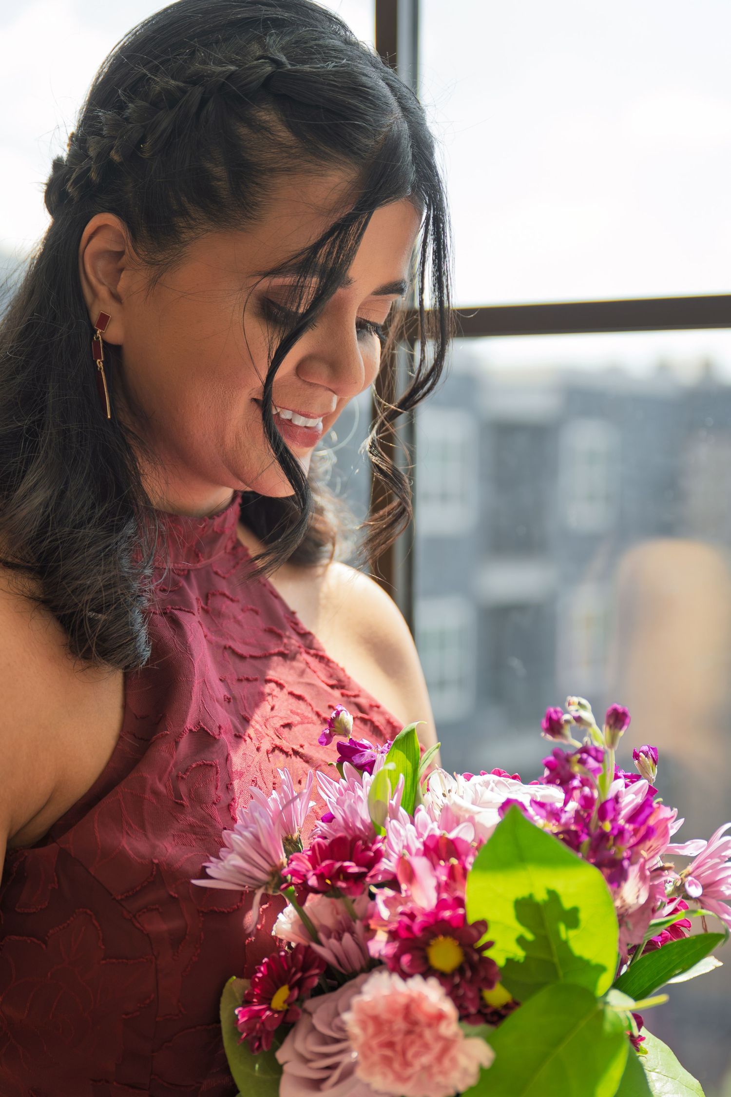 Smiling bride in burgundy dress admiring colorful pink and purple wedding bouquet by window.