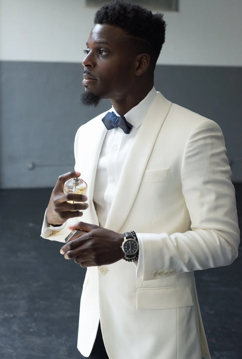 Stylish groom in white blazer and navy bow tie holding fragrance bottle at wedding venue.