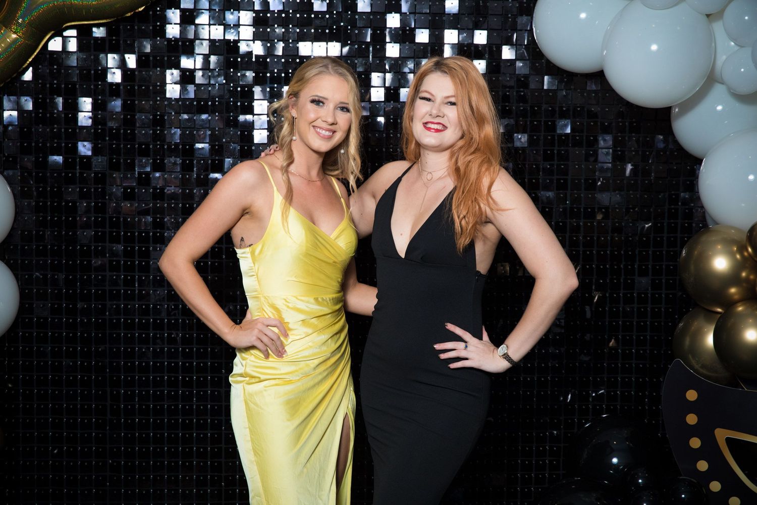 Two women in elegant formal gowns posing in front of a sequined backdrop with balloon decorations.