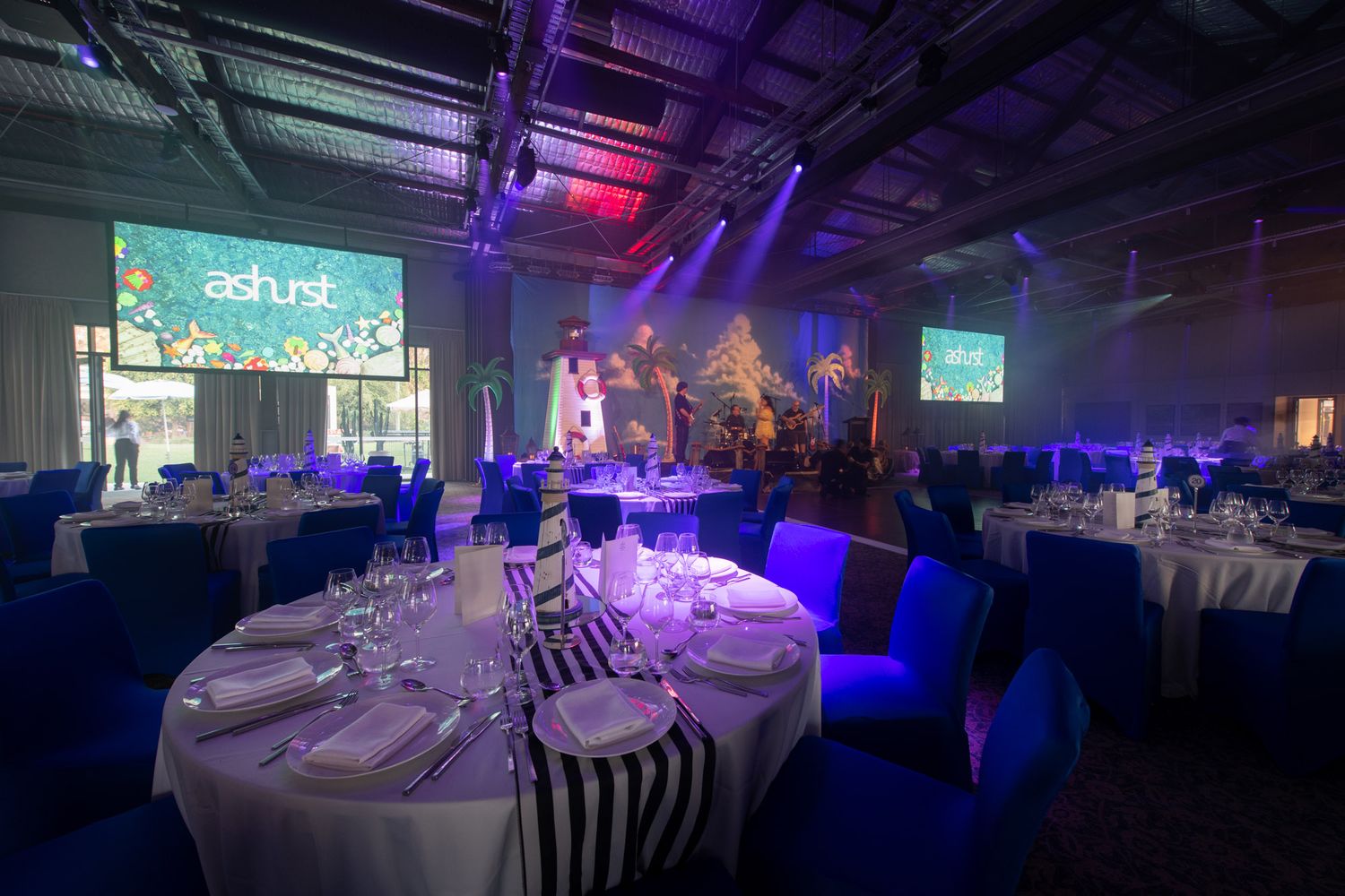 Corporate gala dinner setup with blue lighting, round tables, and Ashurst branded projection screens on stage.
