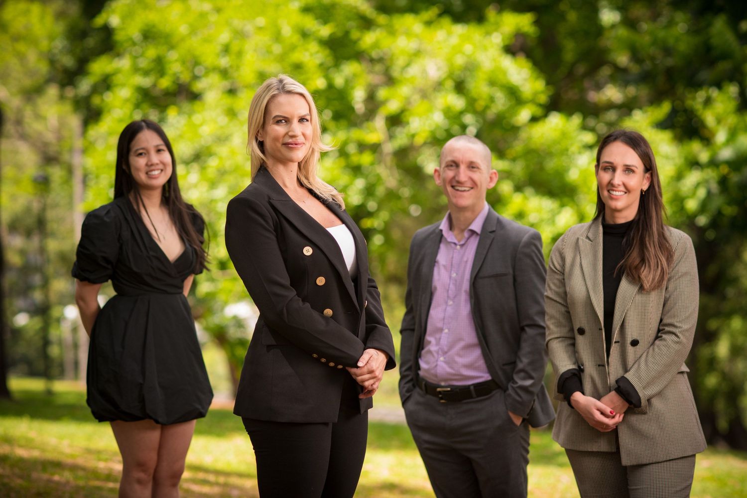 Four business professionals, three women and one man, posing together in a lush green park setting.