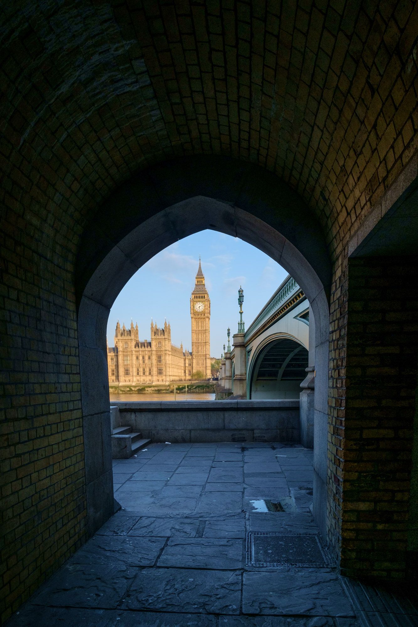 Big Ben and Westminster Bridge framed through a dark brick archway tunnel in London at golden hour.