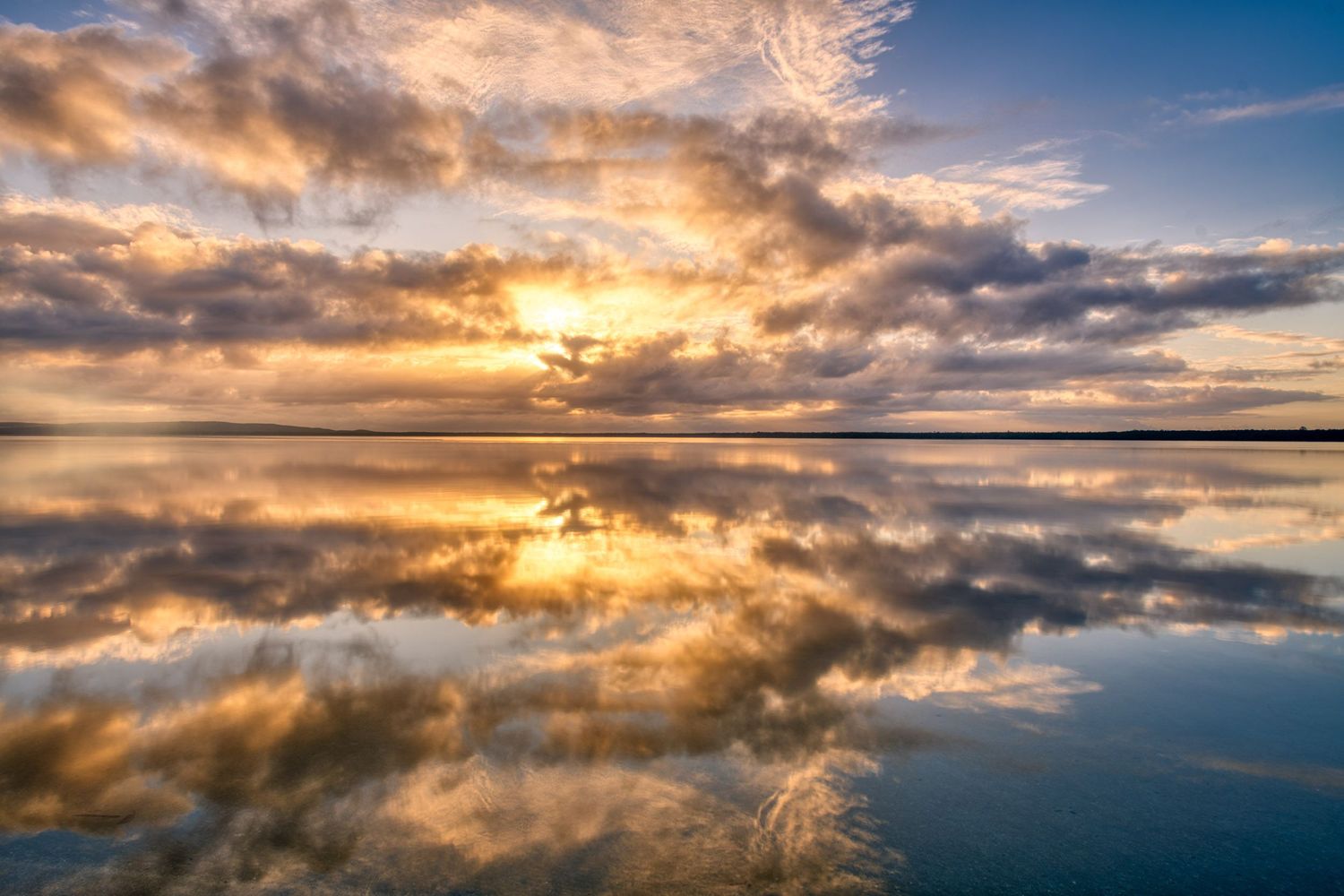 Stunning golden sunrise with dramatic clouds perfectly mirrored on a glassy calm lake surface.