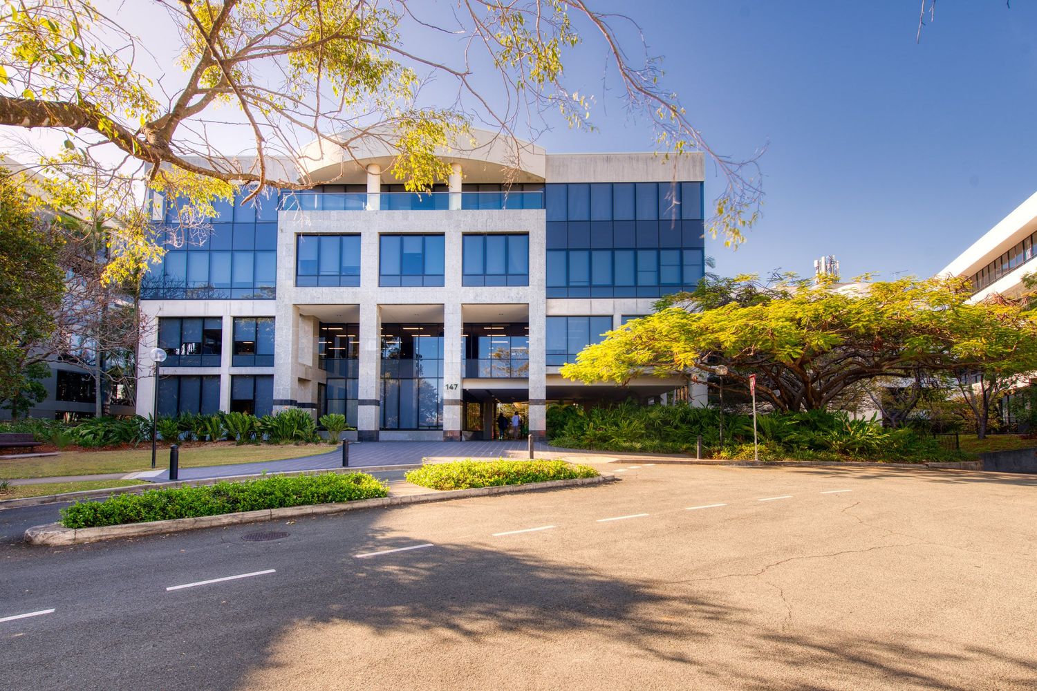 Modern three-story commercial office building with blue glass facade surrounded by manicured gardens and parking area.