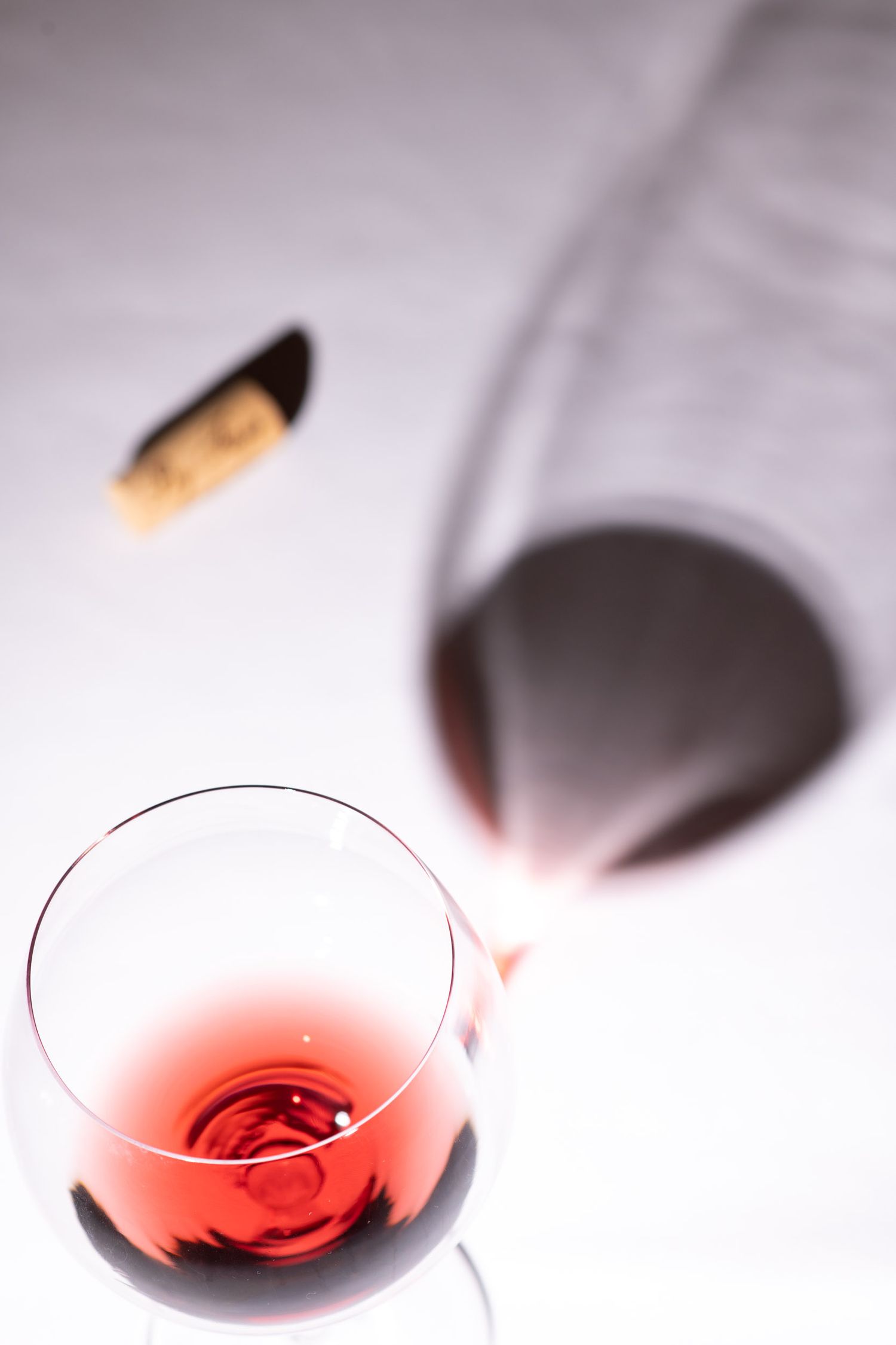 A glass of red wine beside a decanter and cork on a white surface.