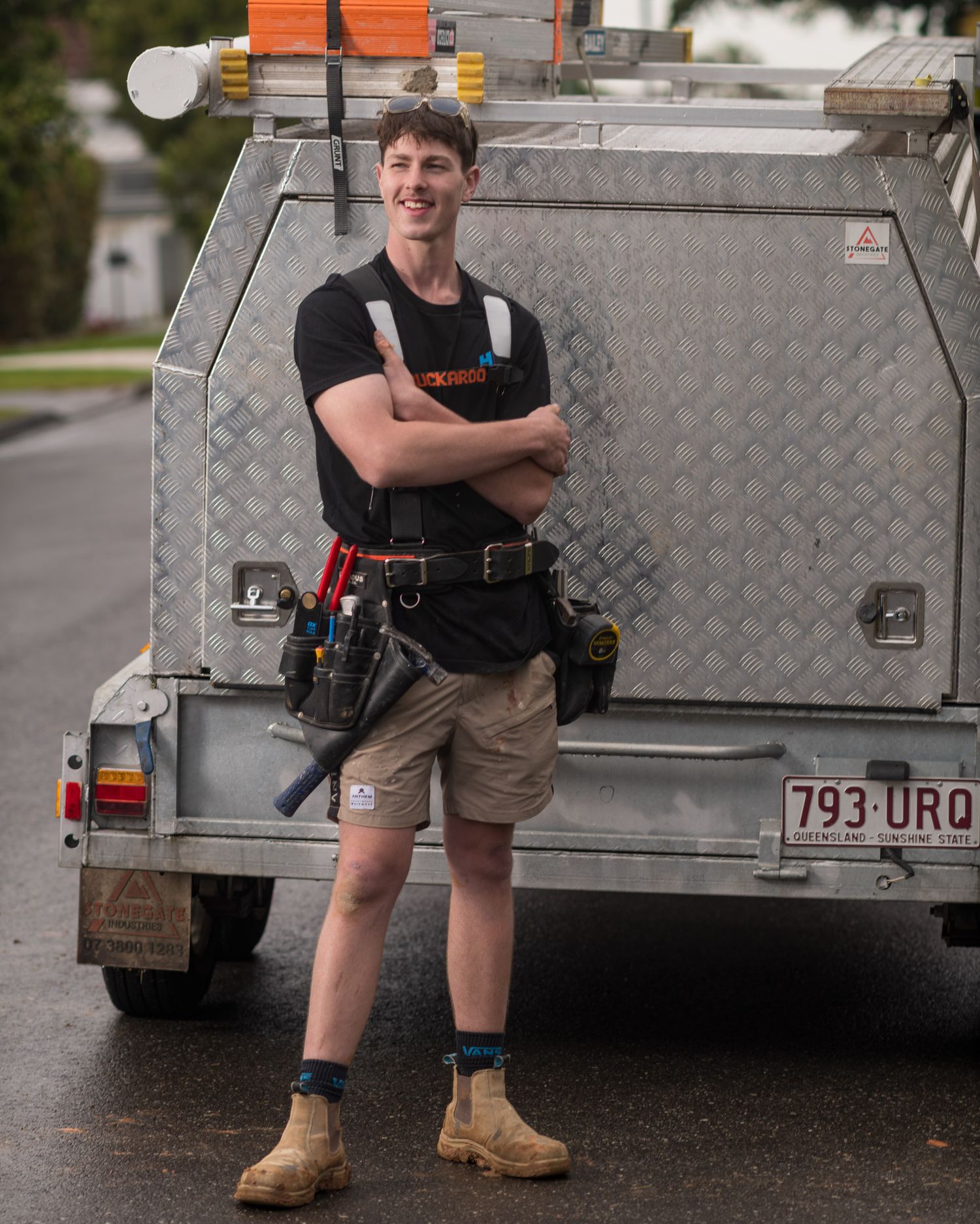 Young male tradesman in work gear smiling with arms crossed beside a utility truck.