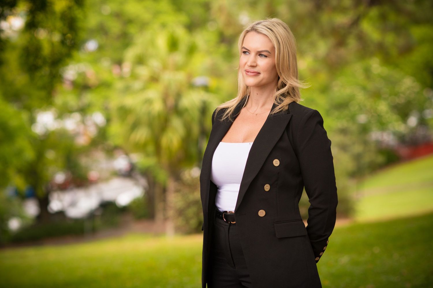 Confident blonde woman in black blazer posing in an outdoor park with lush green foliage background.