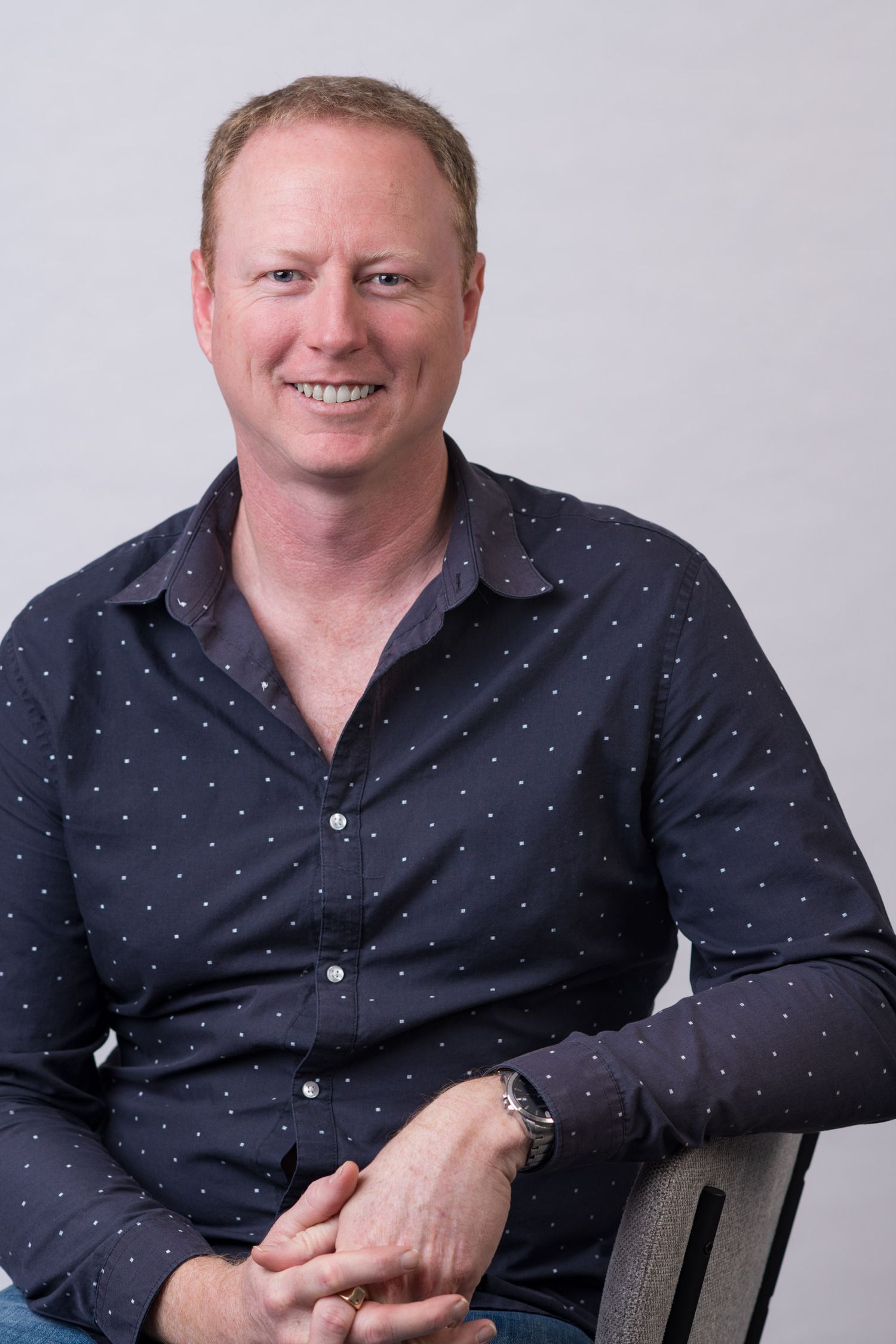 Professional headshot of smiling middle-aged man in dark polka dot shirt seated against grey background.