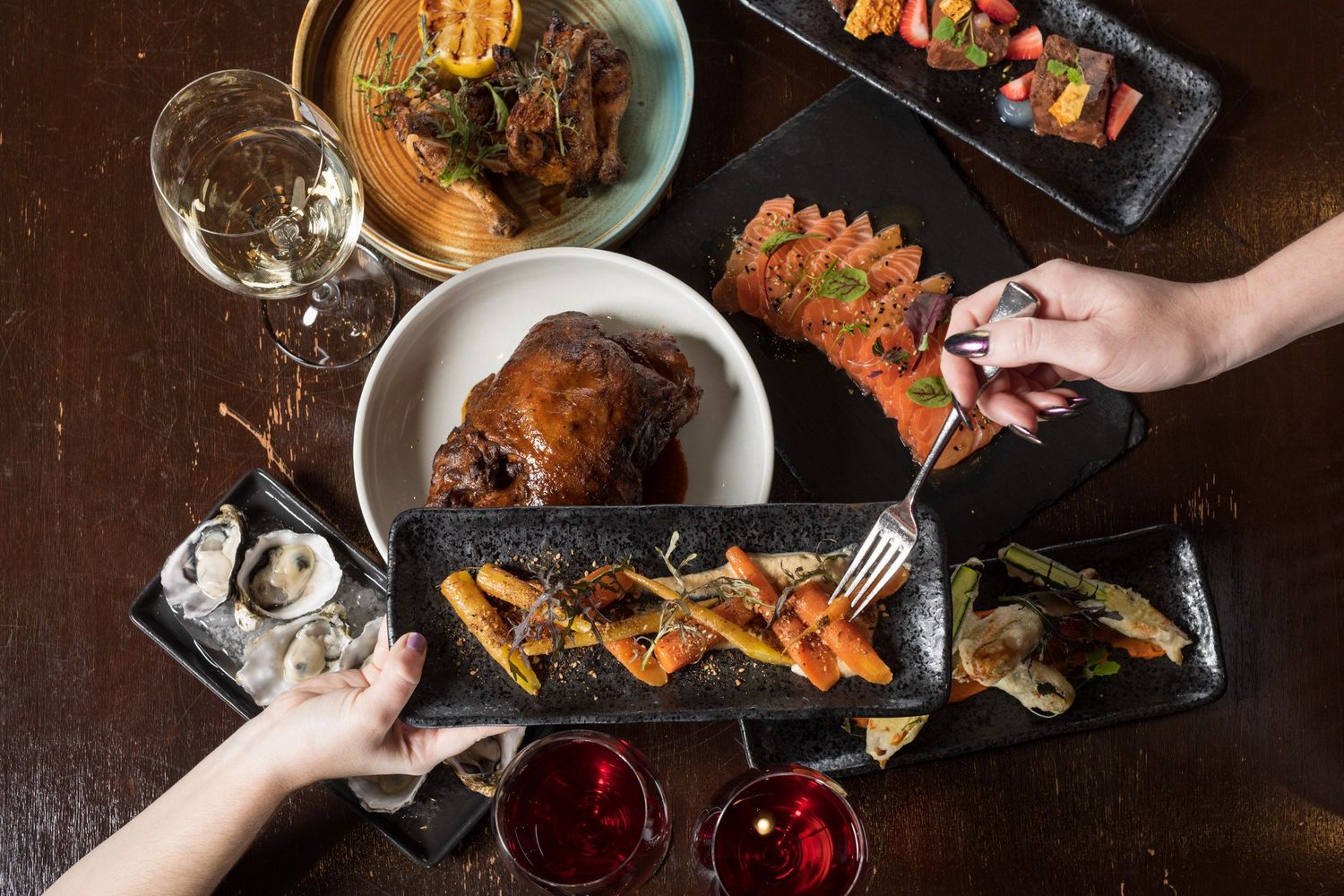 Overhead view of hands sharing multiple gourmet dishes on a dark wooden restaurant table.