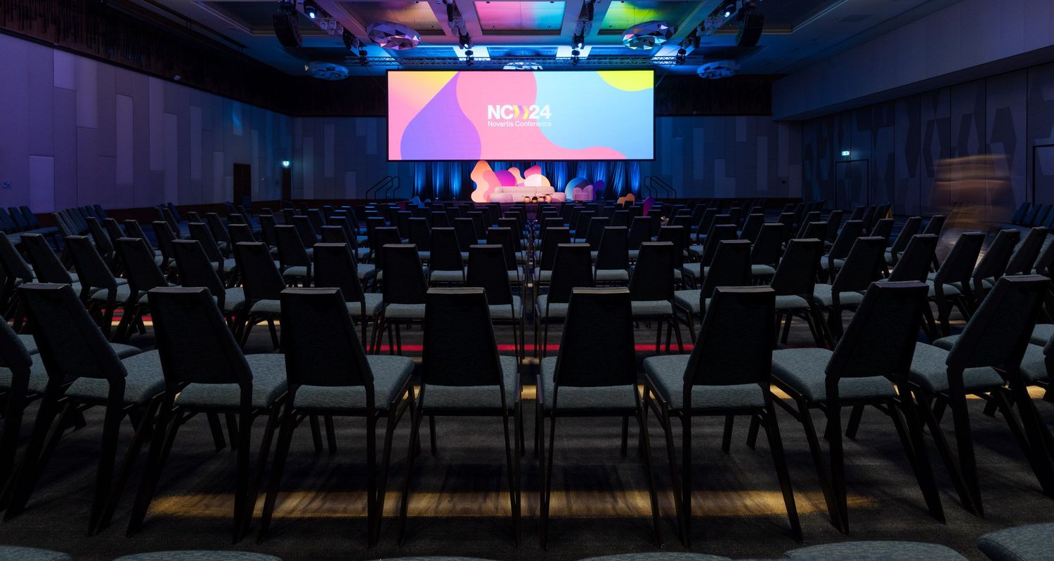 Empty modern conference room with rows of grey chairs facing a bright HD24 branded presentation screen on stage.