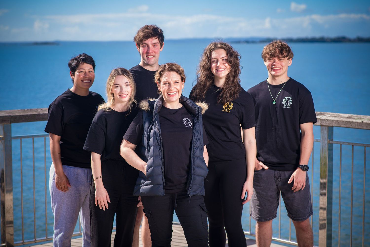 Group of six young people in black shirts smiling together on coastal boardwalk with ocean backdrop.