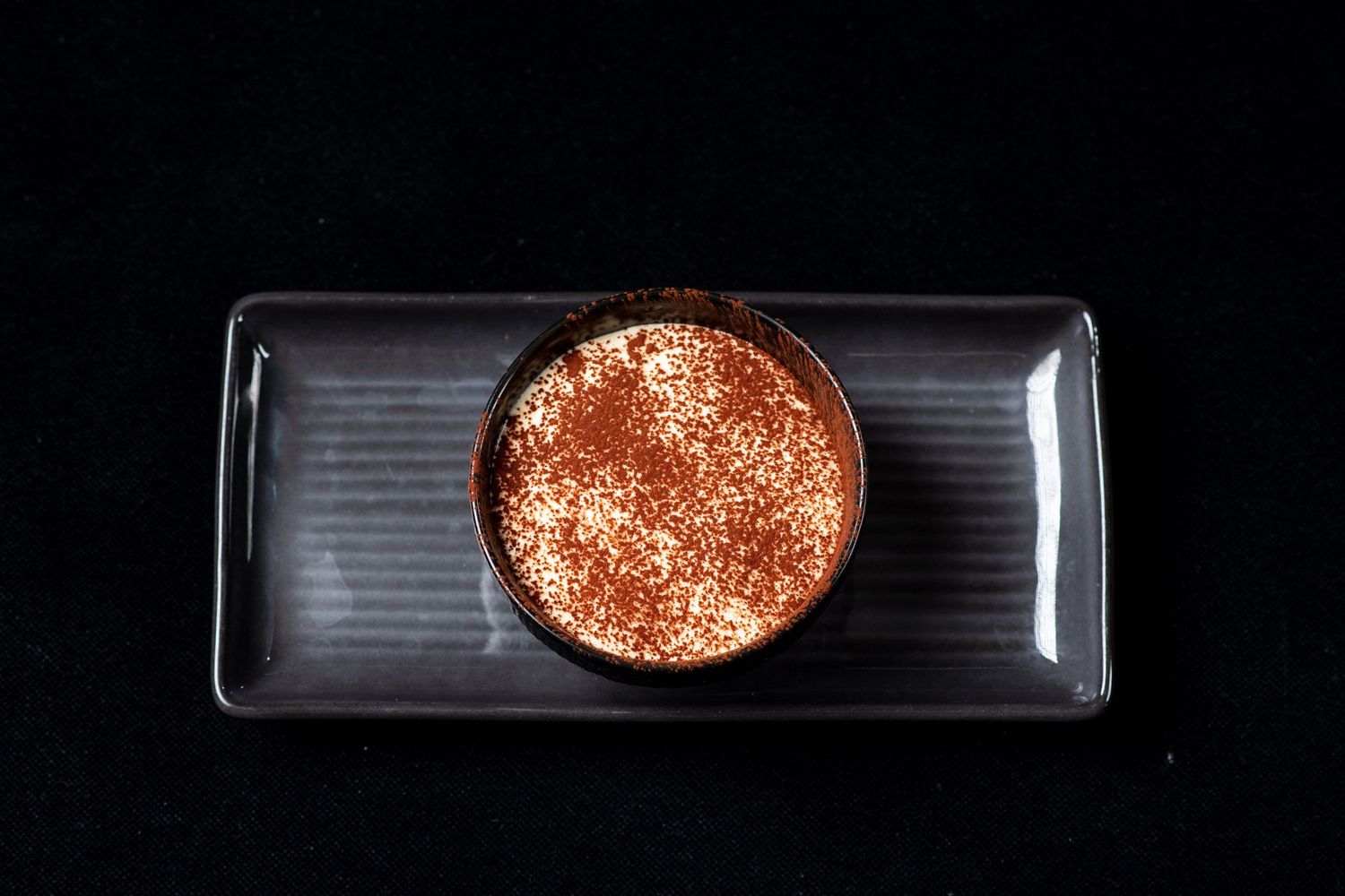 Elegant chocolate dessert dusted with cocoa powder served in a round ramekin on a dark rectangular plate.