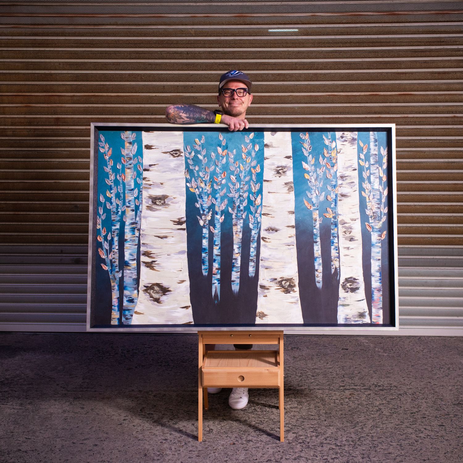 Person in cap peeks over large framed blue birch tree artwork displayed on wooden easel.