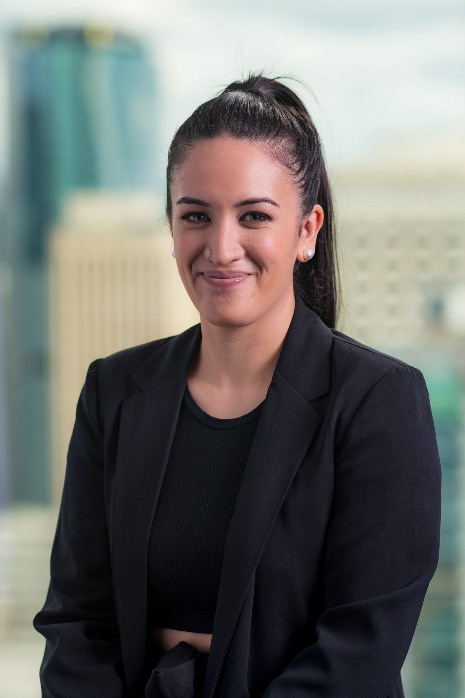 Young woman with dark hair in ponytail wearing black blazer with city buildings in the background.