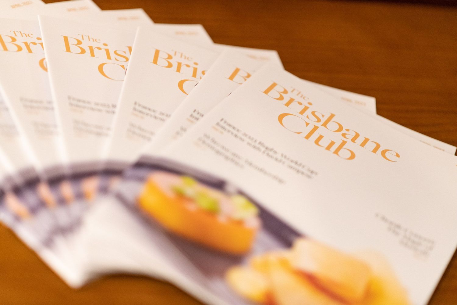 Fanned display of The Brisbane Club magazines featuring gourmet food photography on covers.