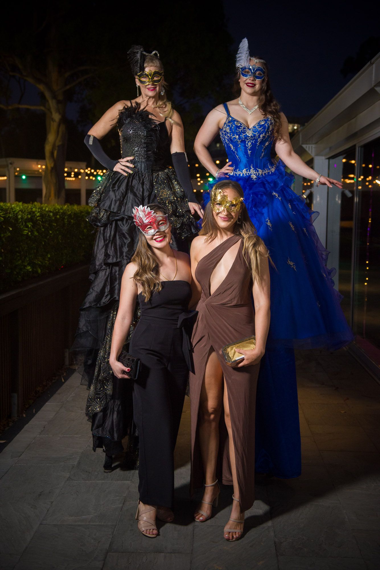 Four women in elegant gowns and masquerade masks pose at a glamorous masquerade ball event at night.