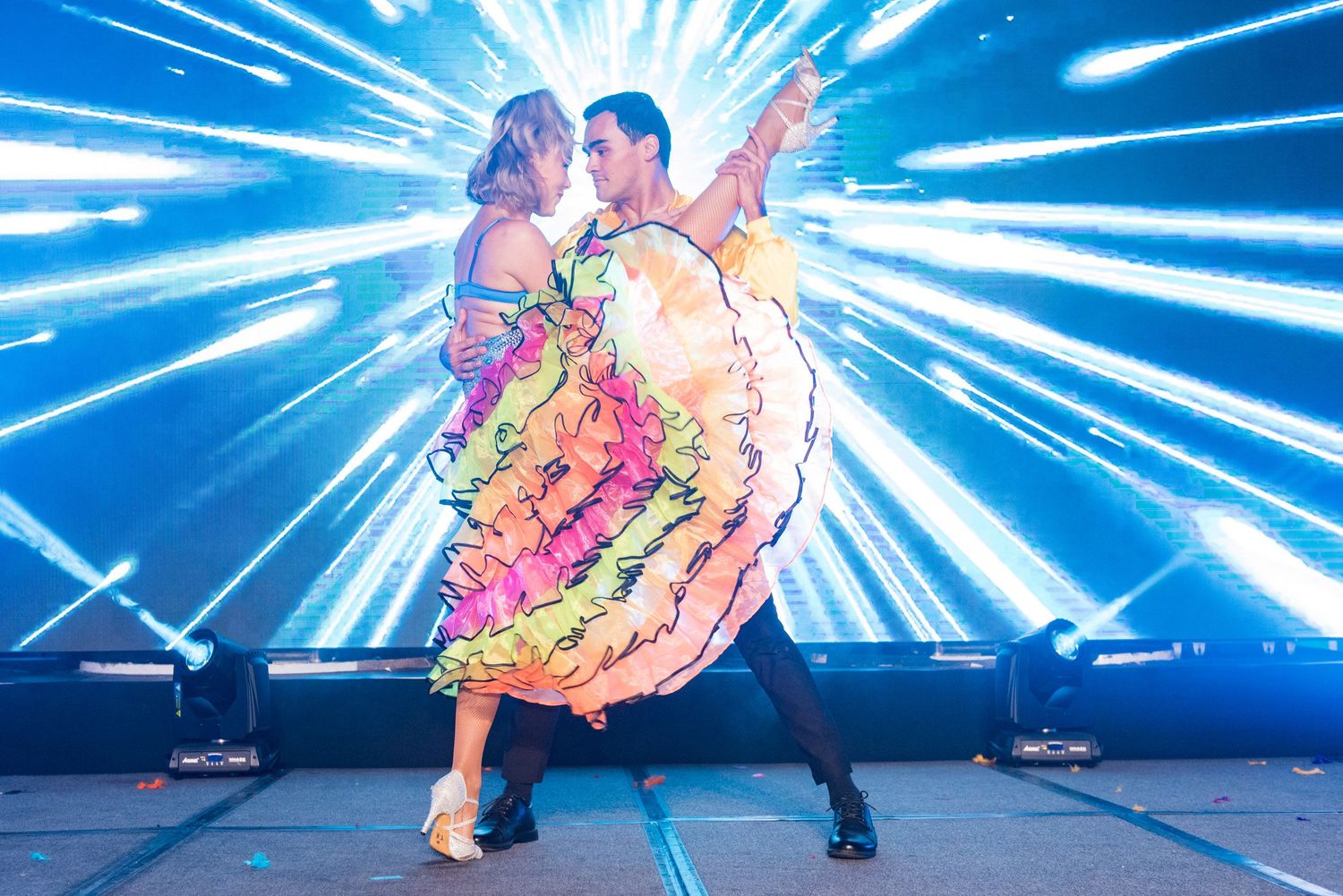 Couple performing elegant ballroom dance on stage with colorful dress and dramatic blue light rays backdrop.