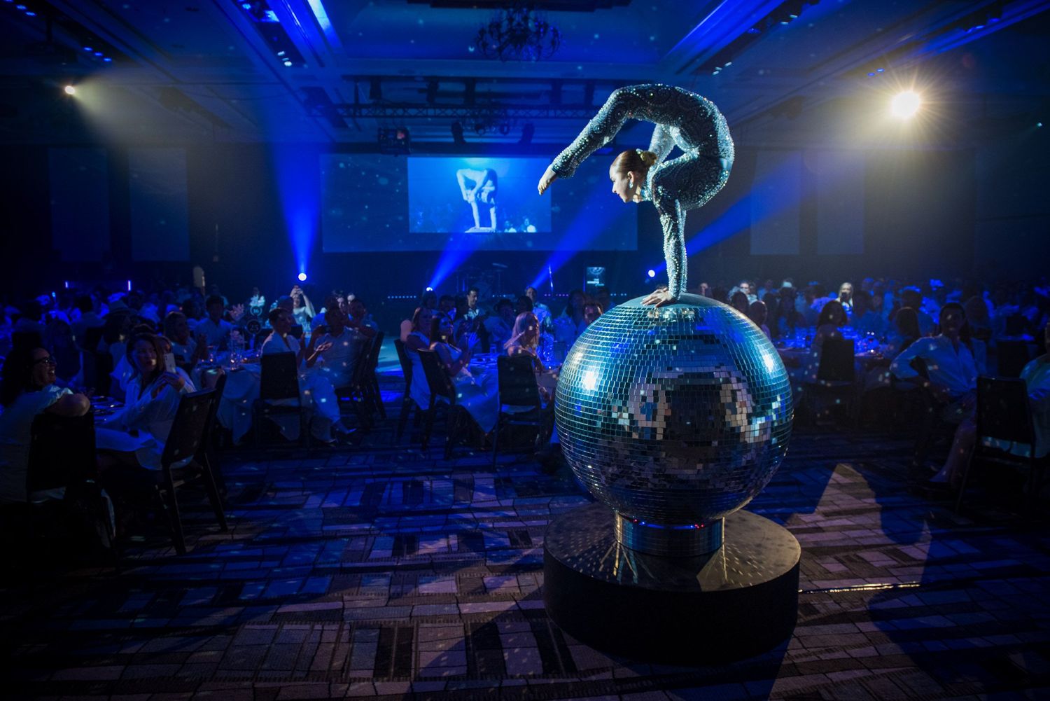 Acrobat balancing on disco ball centerpiece at blue-lit corporate gala dinner.